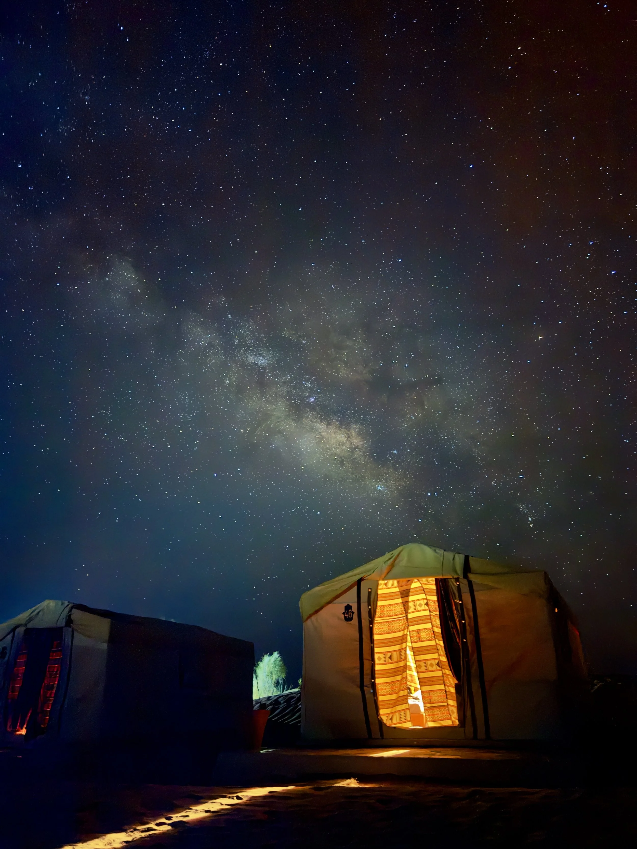 Night sky filled with stars and the Milky Way galaxy above a lit-up tent in a desert landscape.