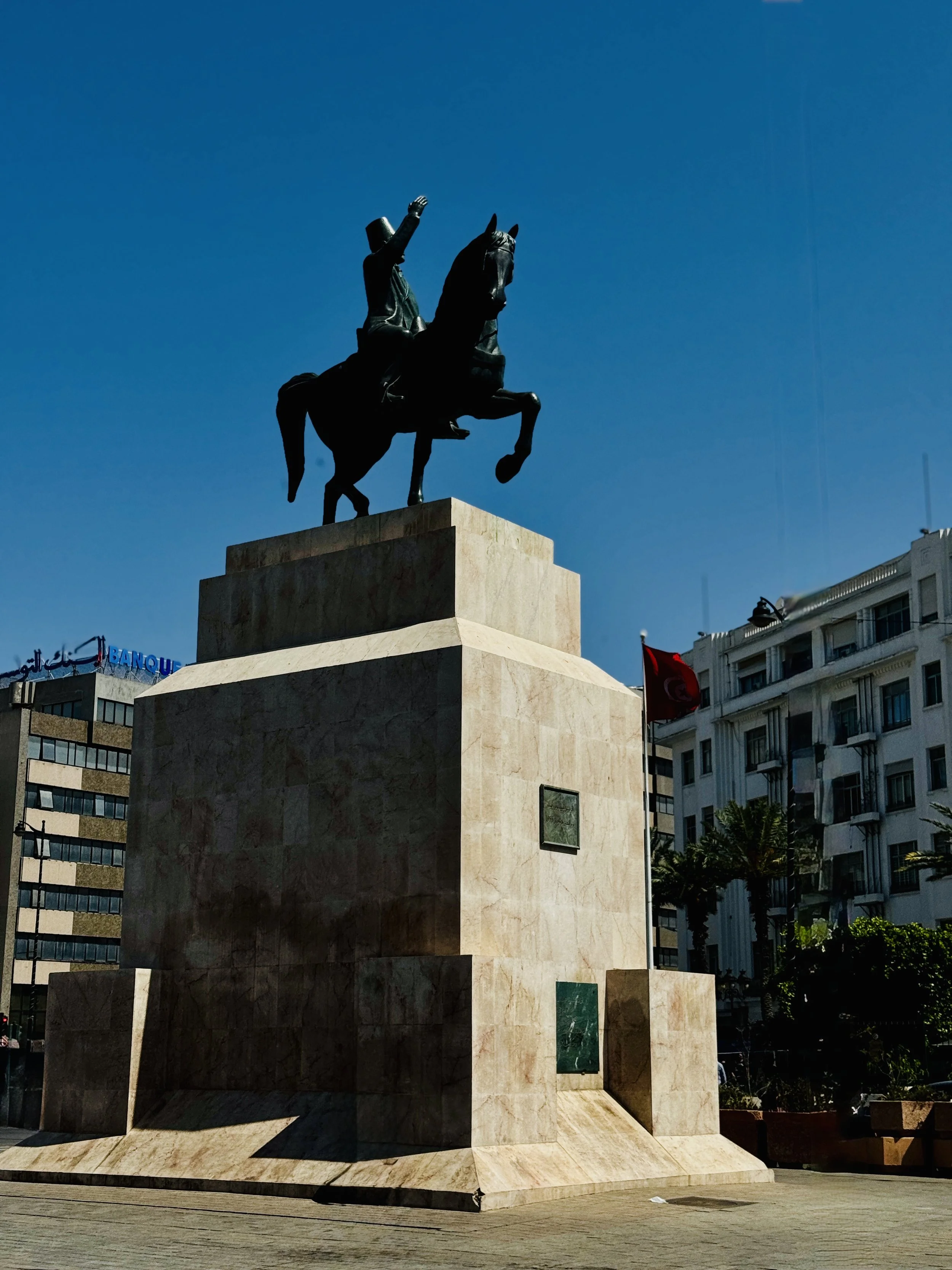 A bronze equestrian statue of a man in a hat, mounted on a horse, is situated on a large beige stone pedestal in an urban setting. Surrounding buildings include modern and classical architecture, with a visible sign reading "BANQUE" in French and other signage, and a Tunisian flag flying.