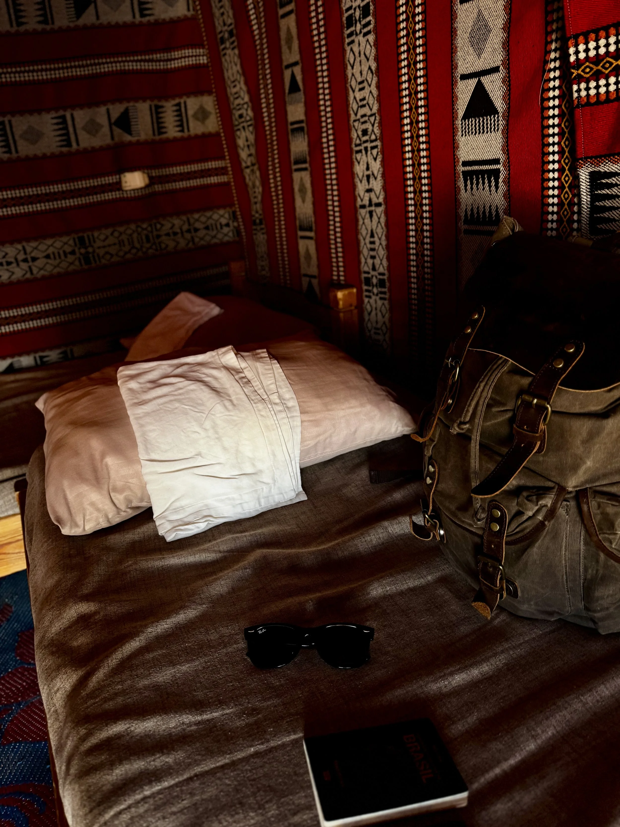 A bed covered with a dark brown blanket, on which a white pillow is placed. Beside the bed, there's a bag with straps, sunglasses, and a book titled 'BRAZIL'. The background features a wall with red, black, and white patterned fabric or wallpaper with geometric designs.