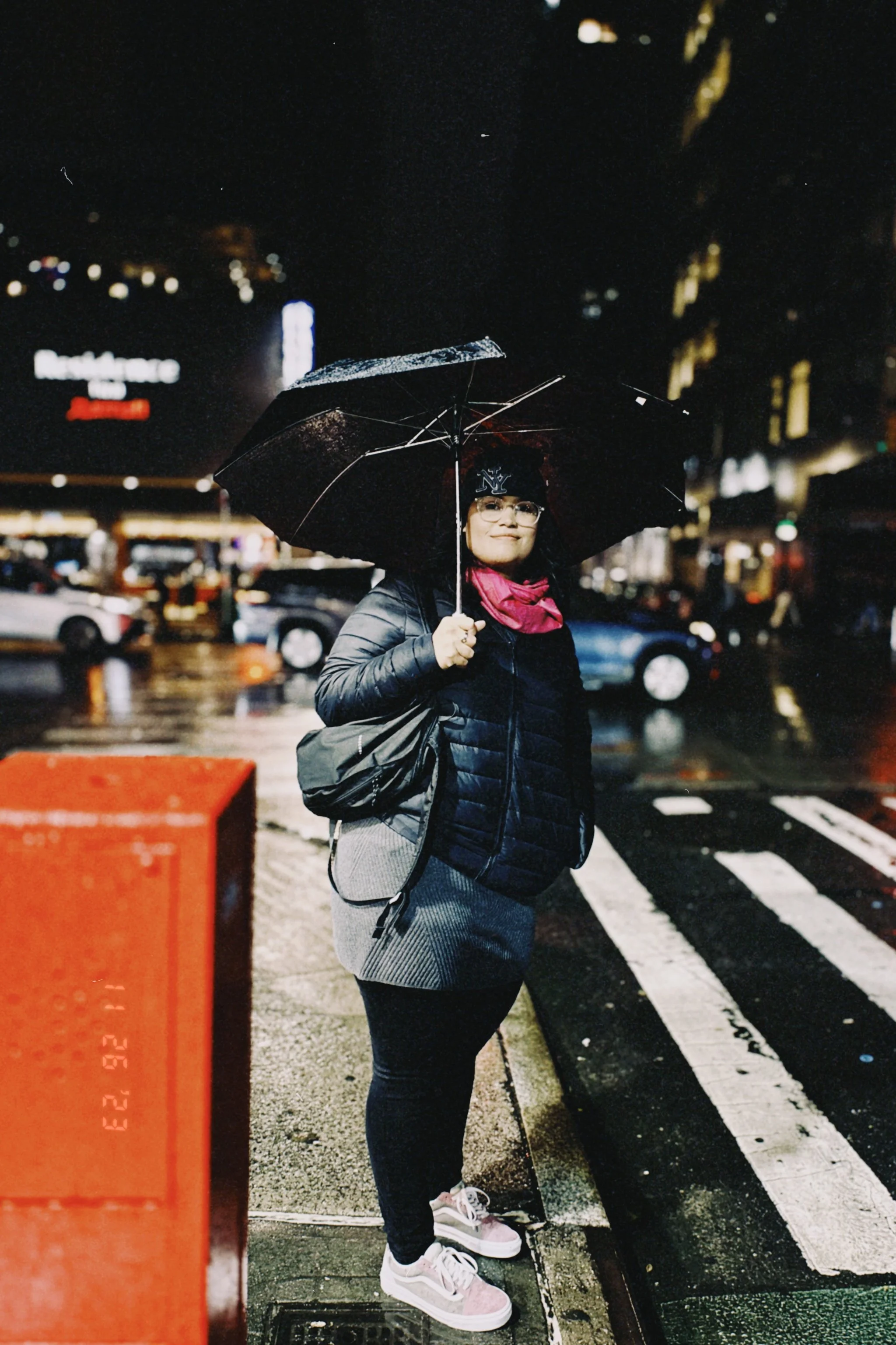 A woman standing on a city street at night, holding an umbrella, wearing glasses, a black jacket, pink scarf, gray pants, and pink and white sneakers, with blurred city lights and cars in the background.