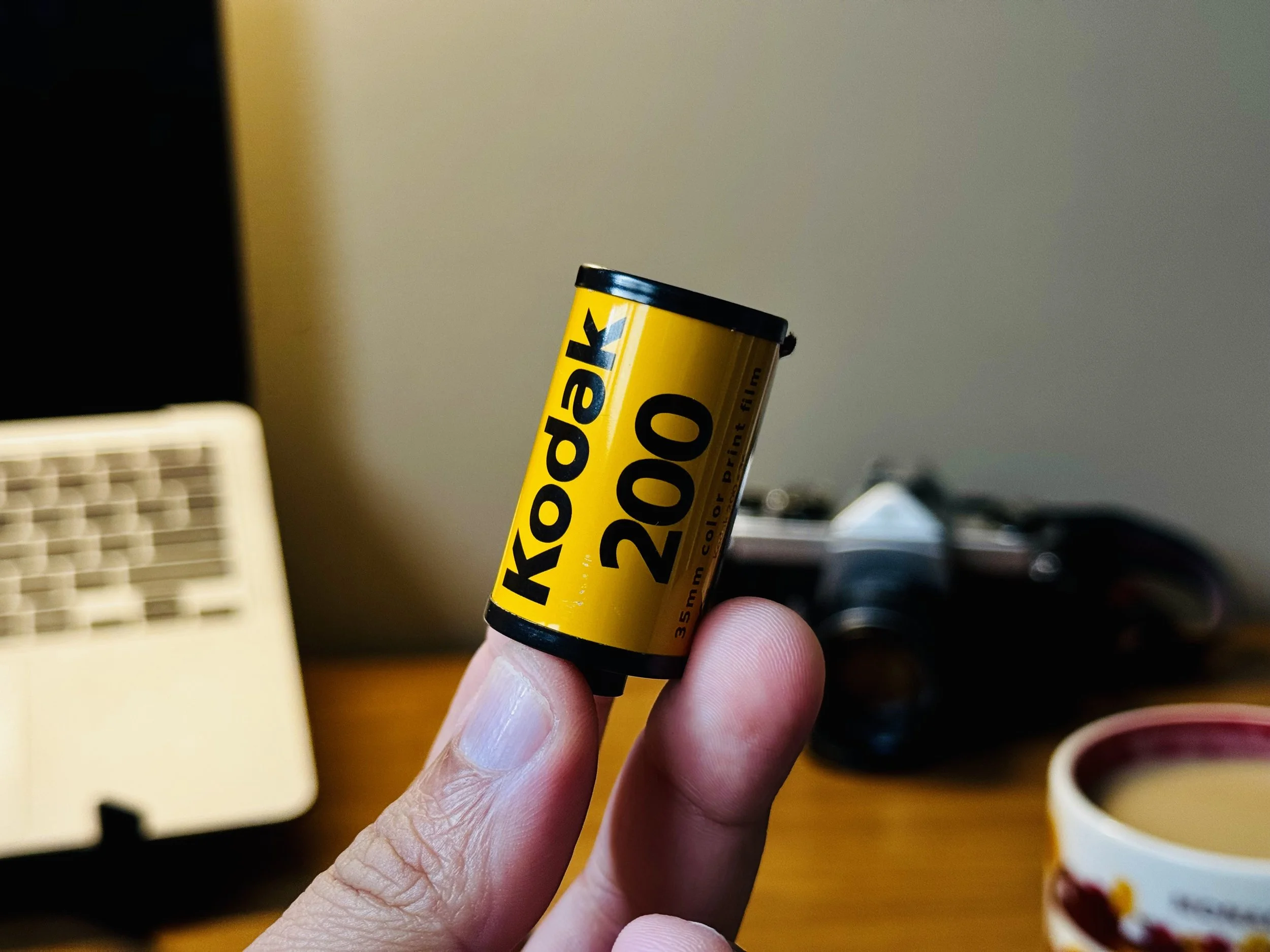 Close-up of a hand holding a yellow Kodak 200 film roll with black text, in a room with blurred background objects.