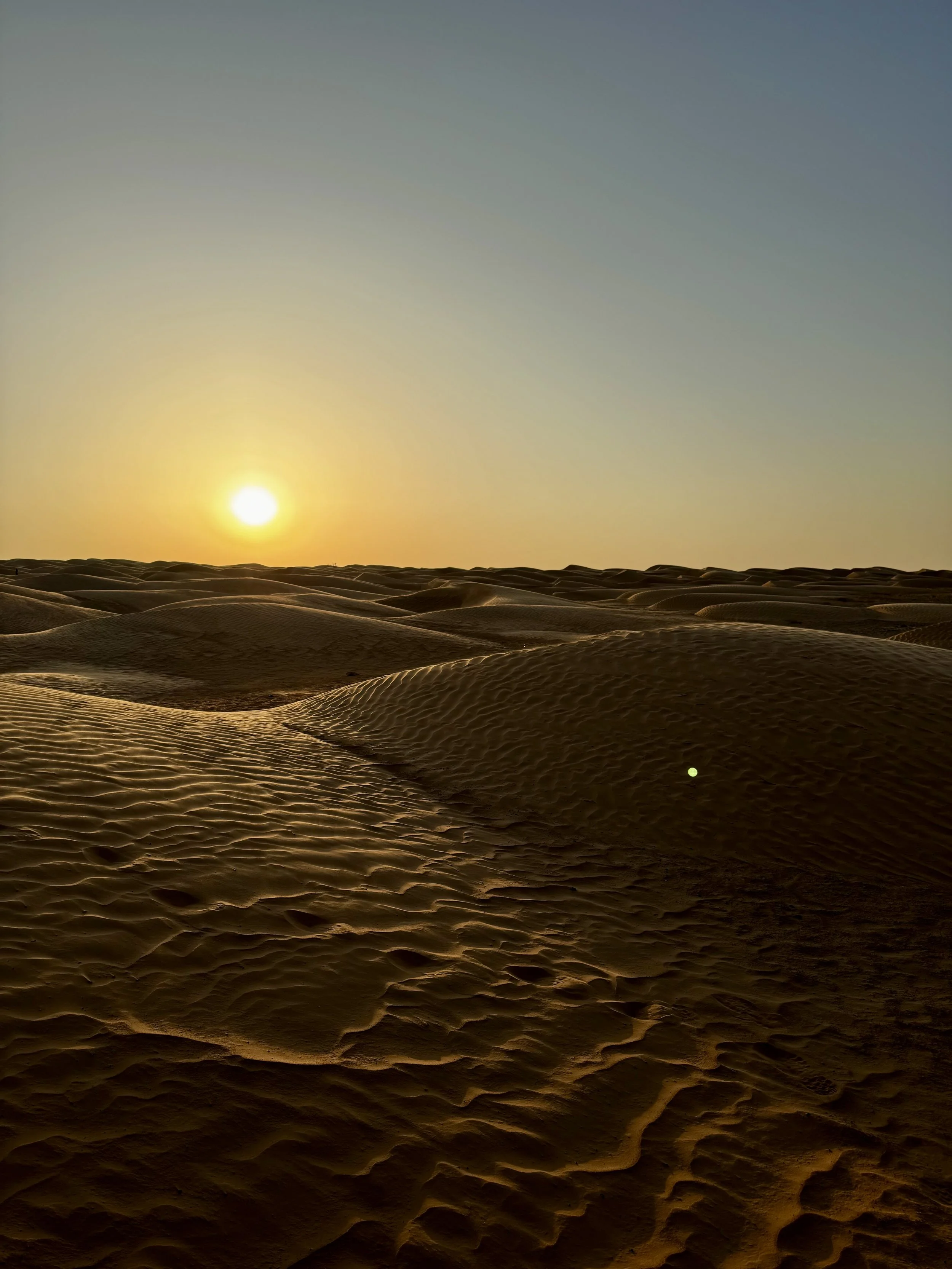 Sunset over a sandy desert with rolling dunes and a clear sky.