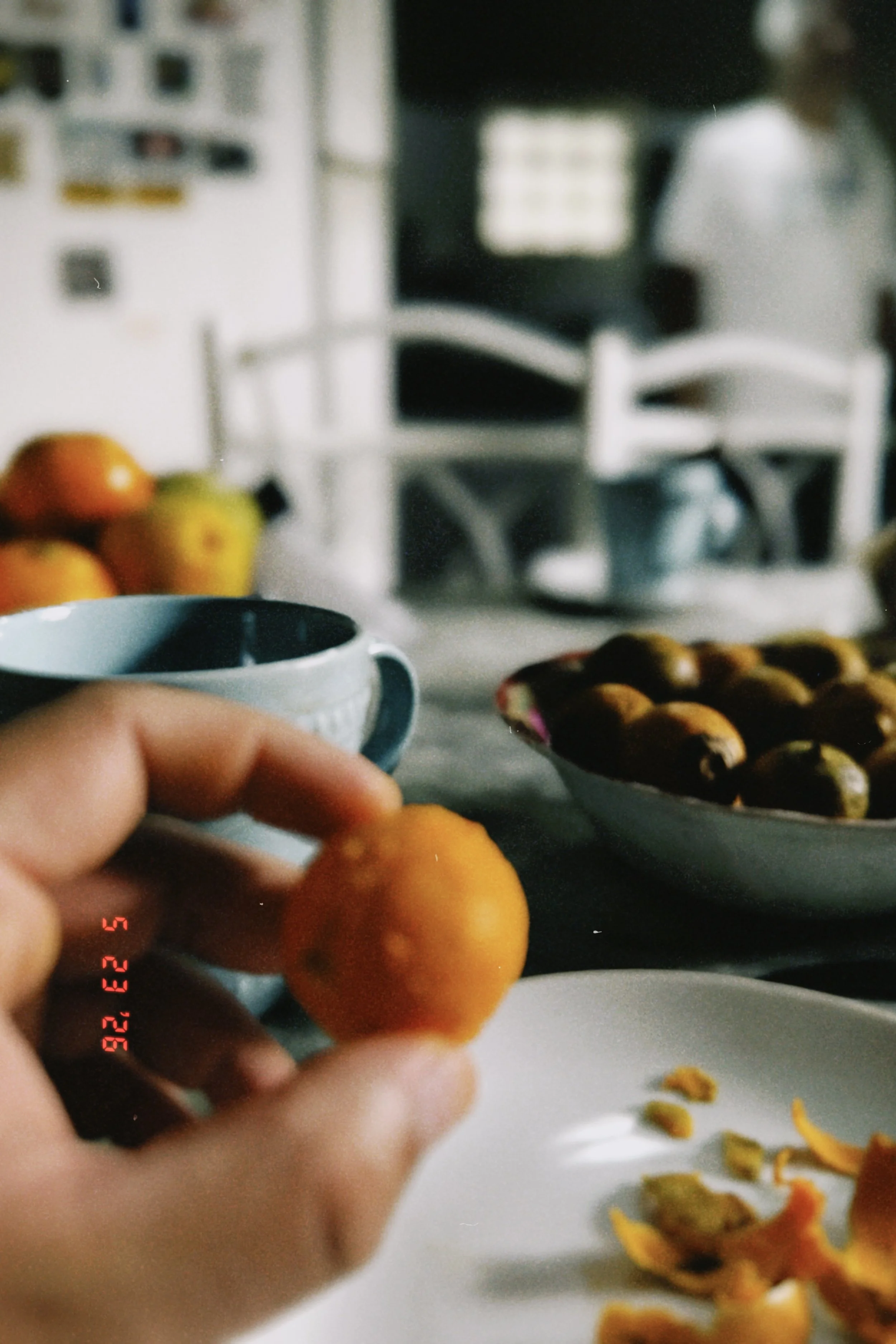 A person holding a small orange fruit, possibly a kumquat, over a white plate of assorted pastries or cookies. In the background, a kitchen or dining area features a white chair, a white basket with more of the same fruit, and blurred kitchen applian