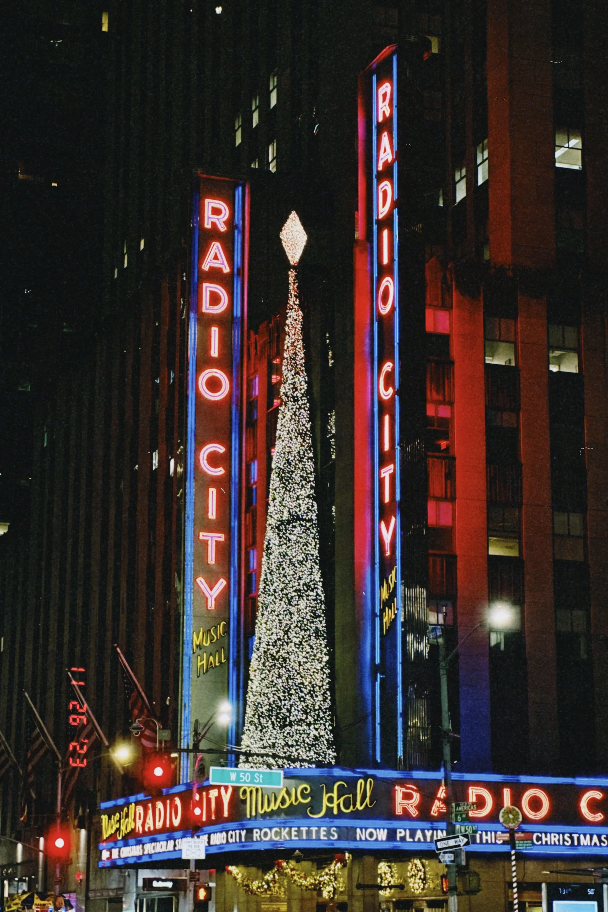 Night view of Radio City Music Hall with bright neon signs and a large Christmas tree in front, decorated with white lights, in New York City.