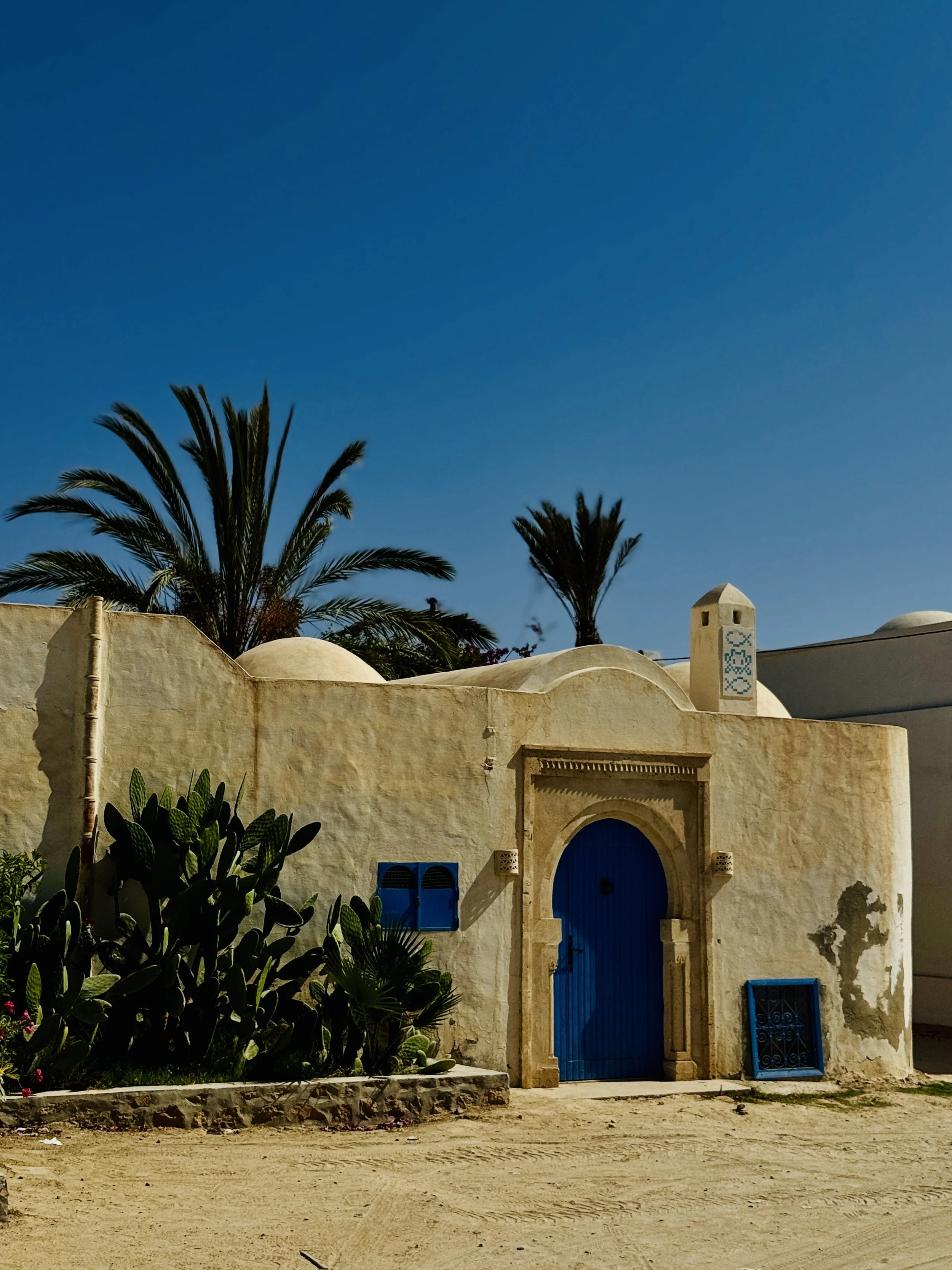 A beige stucco building with a large, arched blue door and small blue window. Tall palm trees and desert plants are in front, under a clear blue sky.