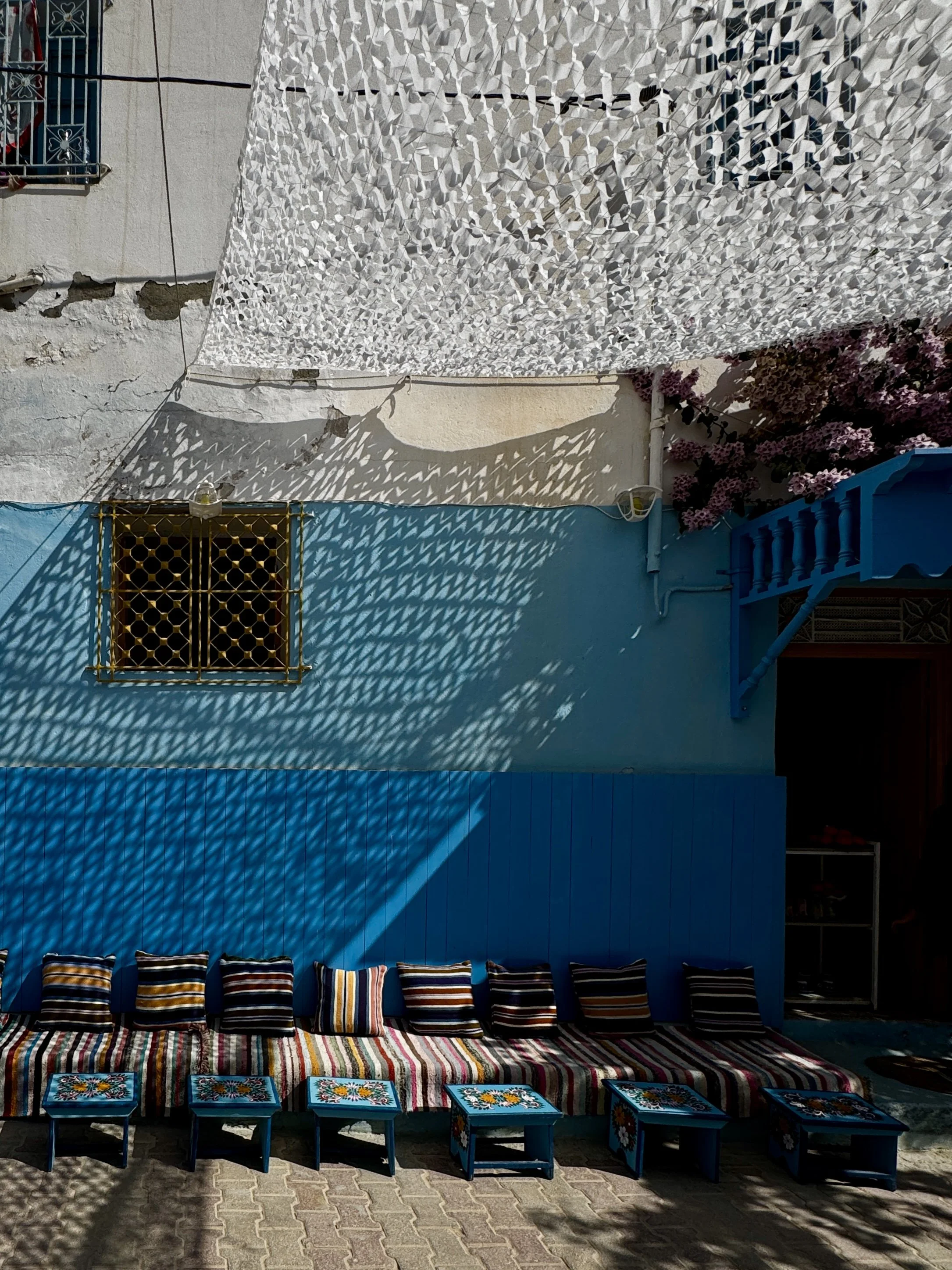 Colorful outdoor seating area with striped cushions, small blue tables with floral designs, a blue wall, a window with a decorative grille, and purple flowering plant, shadow patterns on the wall.