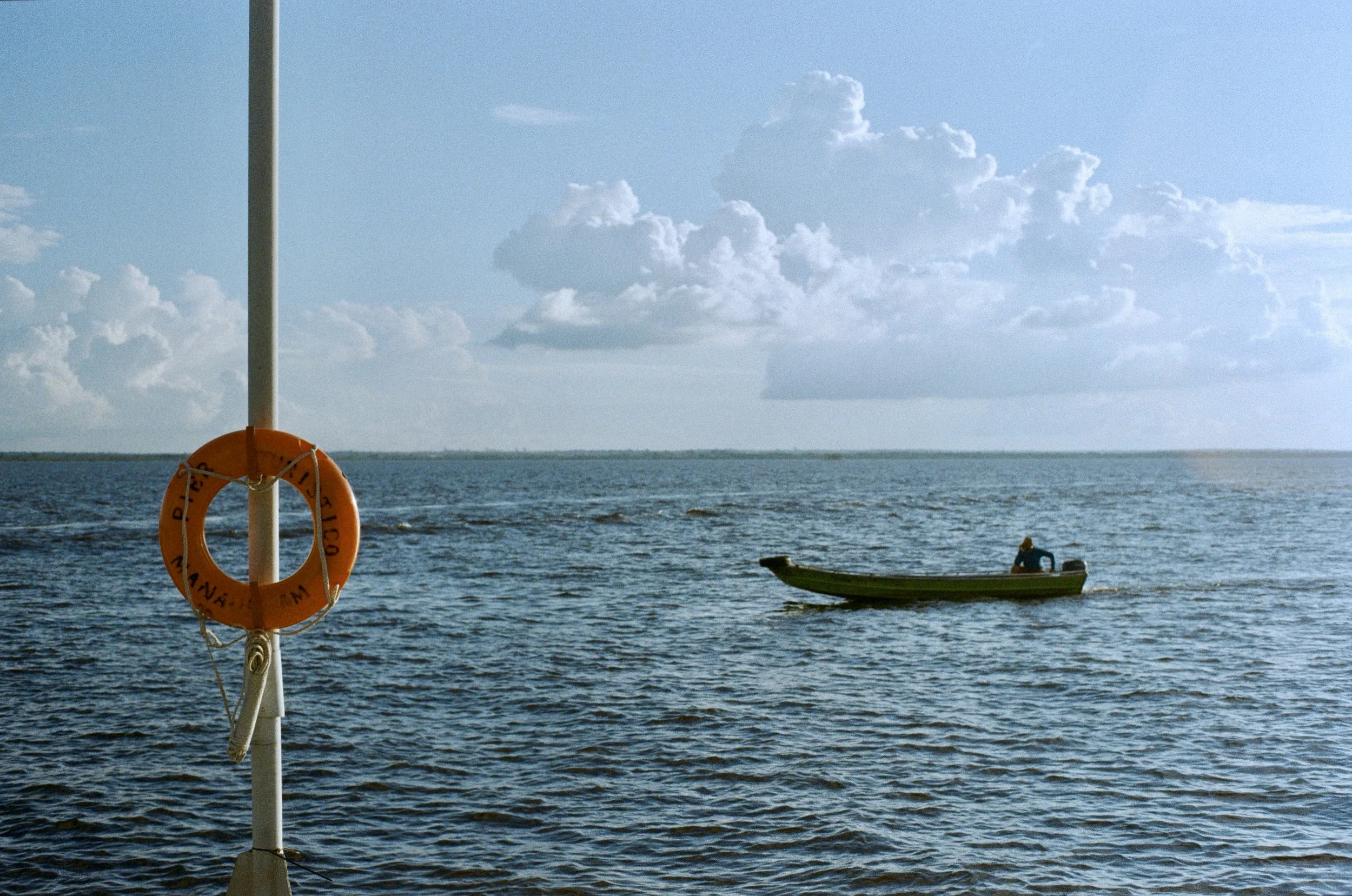 A small boat with a person in it floating on a body of water under partly cloudy sky. In the foreground, part of a boat's pole with an orange life preserver hanging on it.