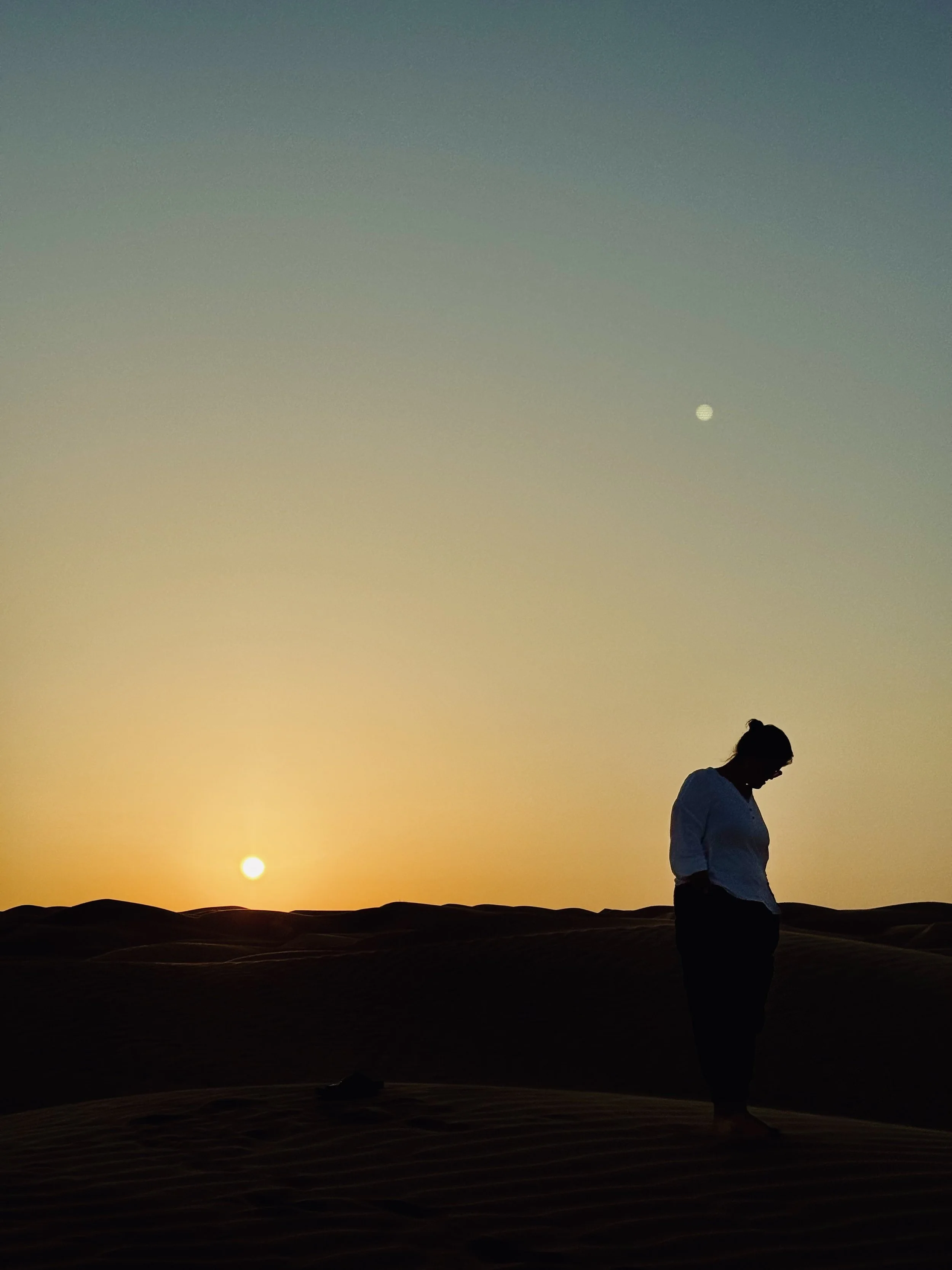 A person standing and looking down in a desert landscape during sunset, with a clear sky, the moon visible, and gentle rolling dunes in the foreground.