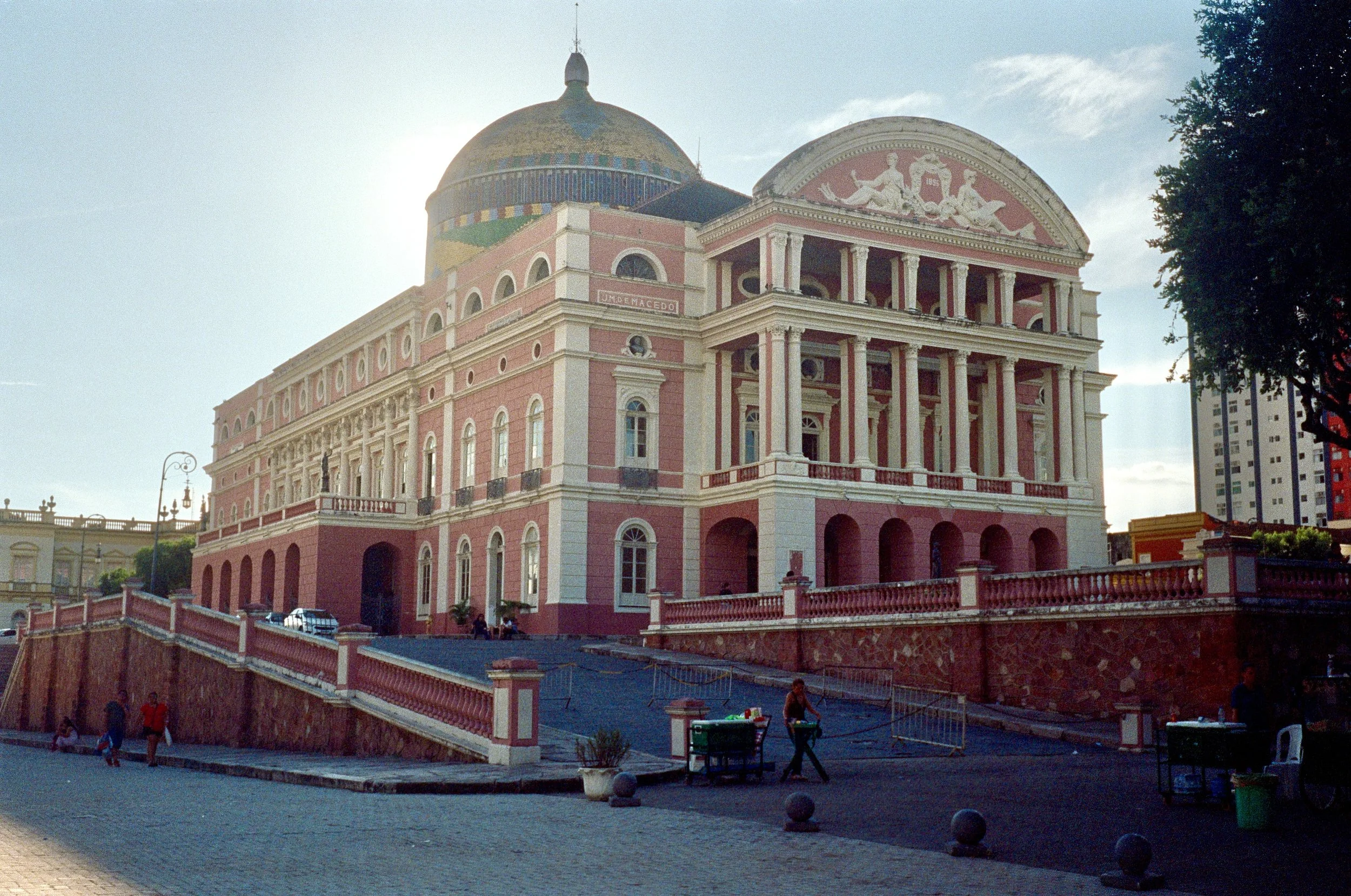 A large, ornate pink and white building with classical architectural features, including columns, a pediment with sculptures, and a domed roof, situated on a street with a sidewalk and few people around.