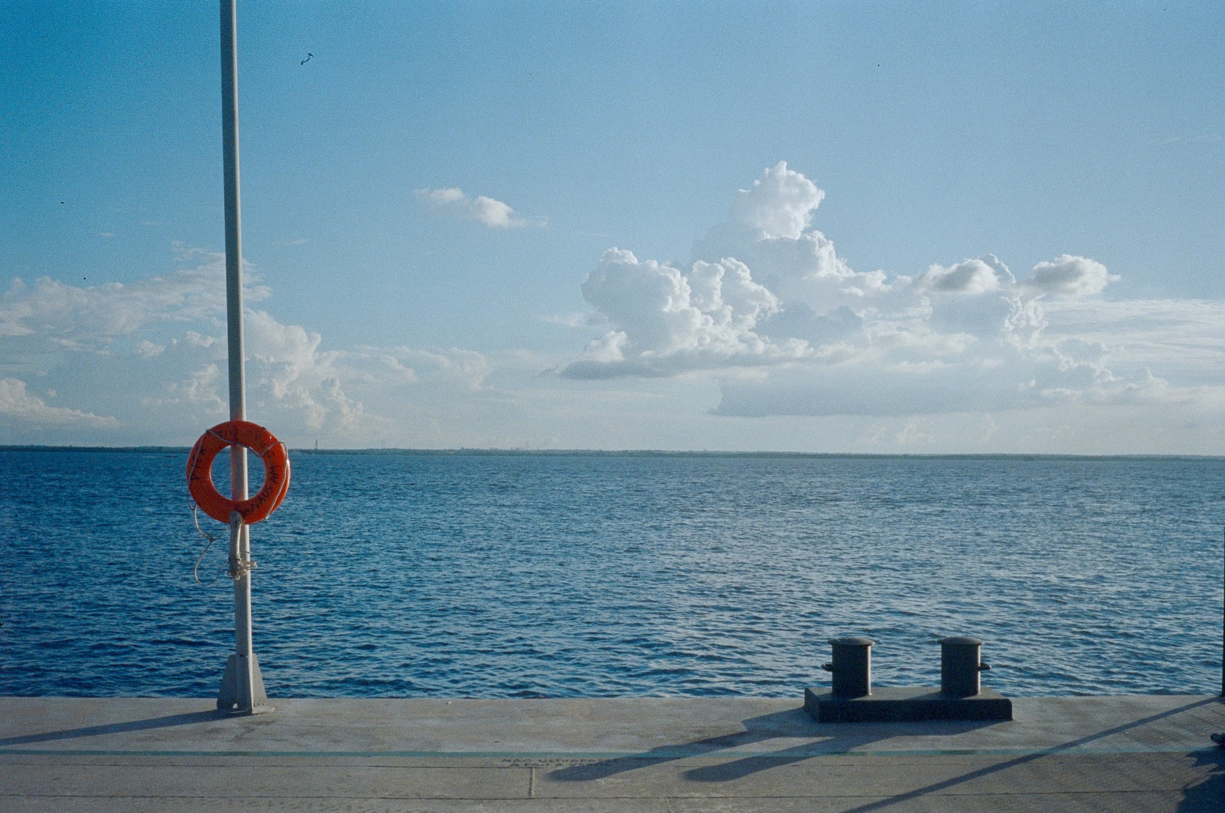 View of a calm blue ocean with a partly cloudy sky and a small boat in the distant horizon. In the foreground, there is part of a boat dock with a white post, a life ring attached to the post, and some bollards on the deck.