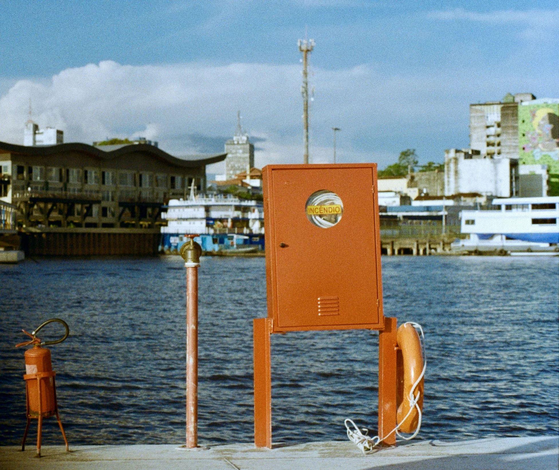 A waterfront scene with a bright orange fire alarm box labeled 'INCENDIO,' a small fire hydrant, and a white coiled cable. In the background, there are buildings, some under construction, with a cloudy sky overhead.