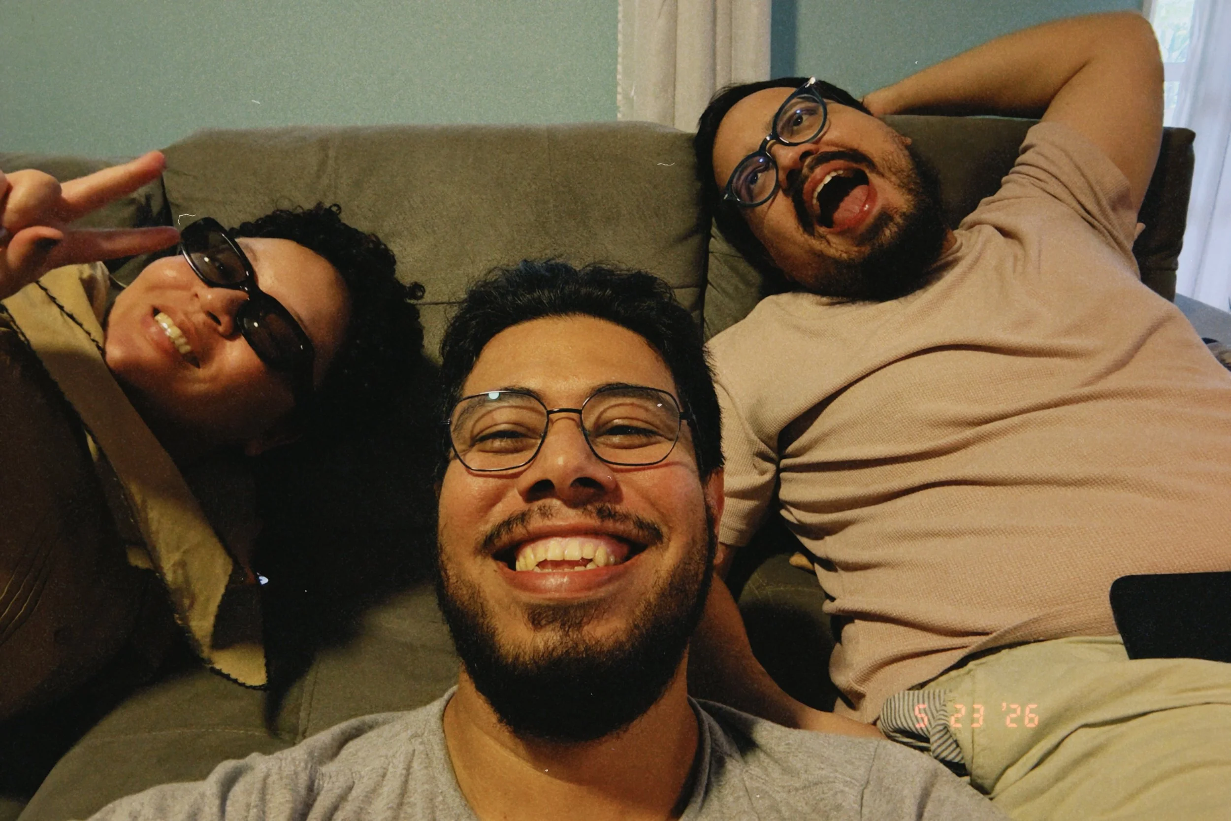 Three people lying on a couch taking a group selfie. They are smiling and appear happy, with one person making a peace sign with their fingers.