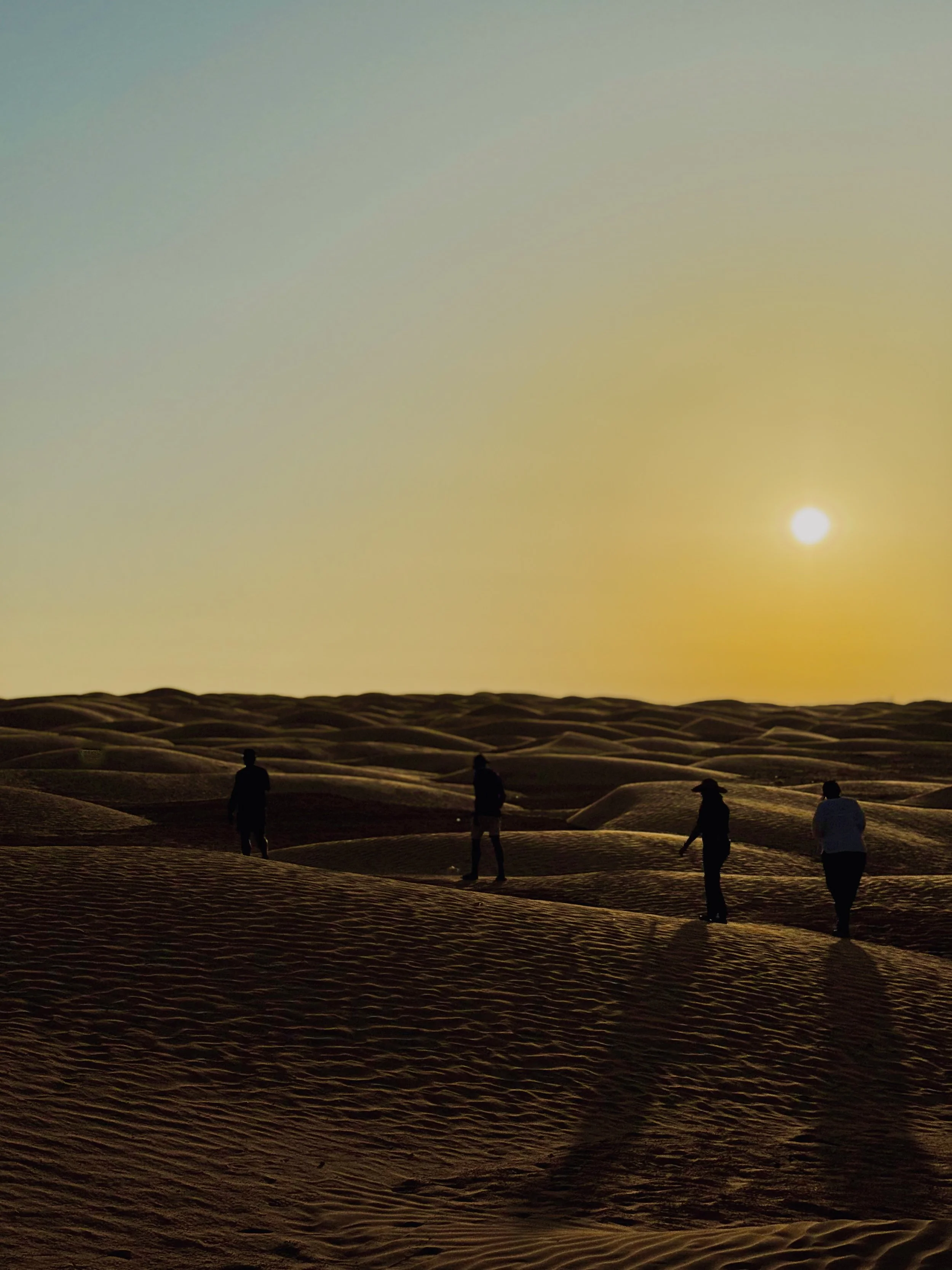 People walking on desert sand dunes at sunset with a bright sun in the sky.