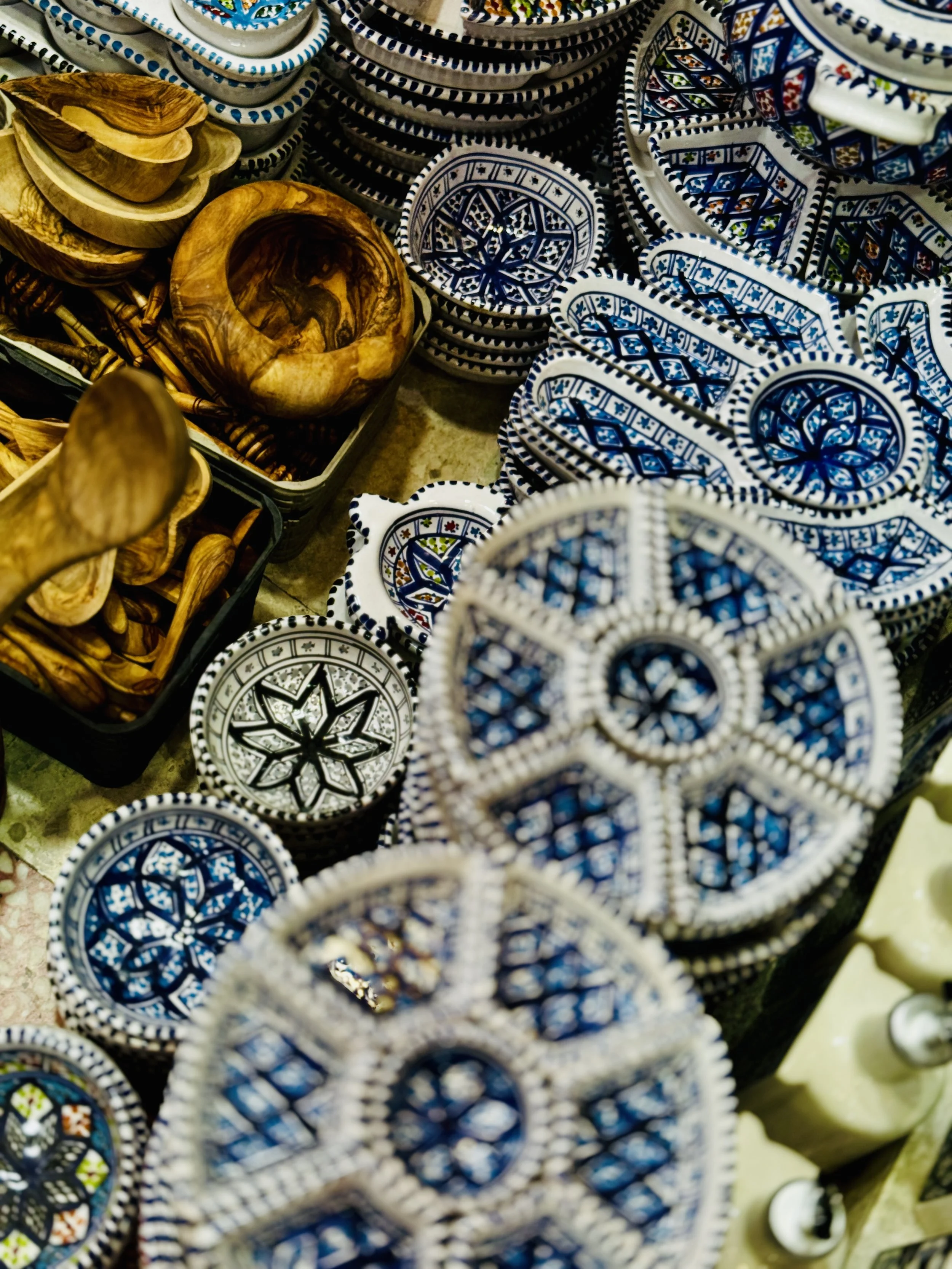 Stacked decorative ceramic bowls with intricate blue, white, and colorful patterns, and wooden bowls, on display at a market.