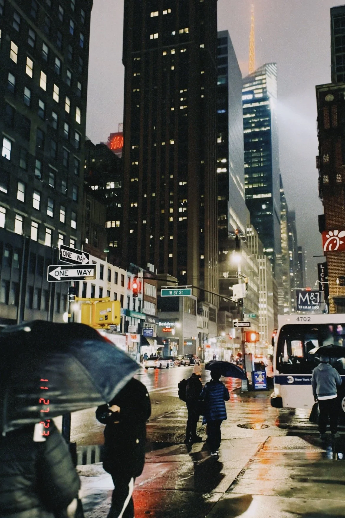 Nighttime city street scene with tall buildings, neon signs, and people walking with umbrellas in the rain.
