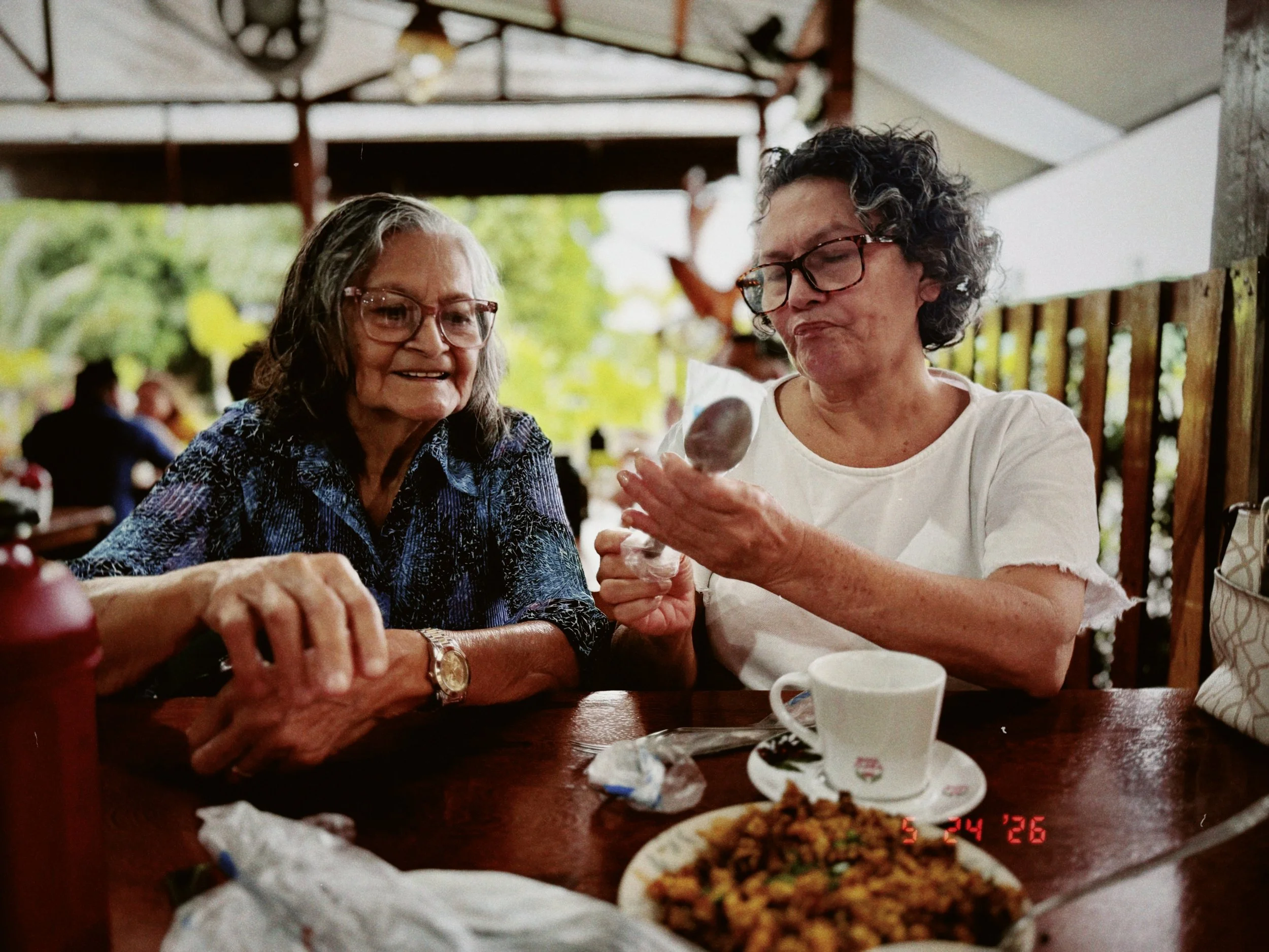 Two elderly women with glasses sitting at a table, enjoying a meal together in a restaurant or cafe. There is food on the table including a bowl of fried rice or similar dish, and a cup.