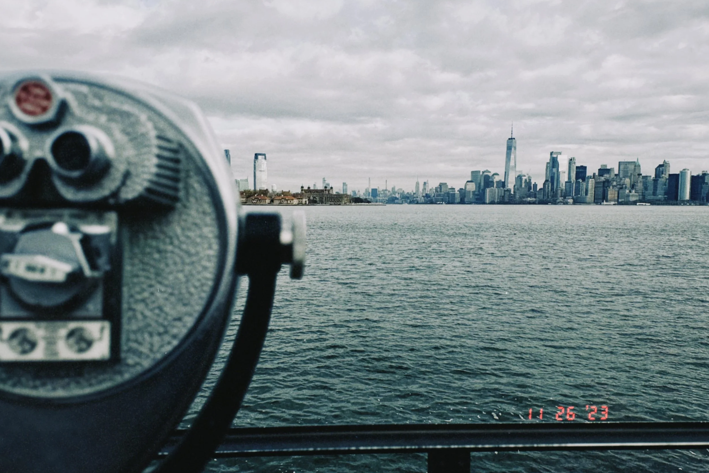 View of New York City skyline seen from across a body of water, with a coin-operated binocular viewer in the foreground, overcast sky.