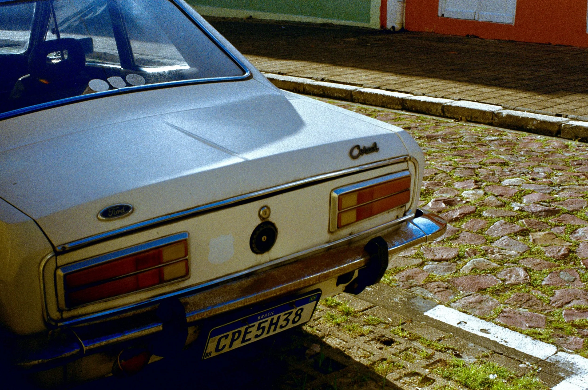 Rear view of a vintage white Ford car parked on a cobblestone street, with a nearby sidewalk and a building with an orange door and green wall.