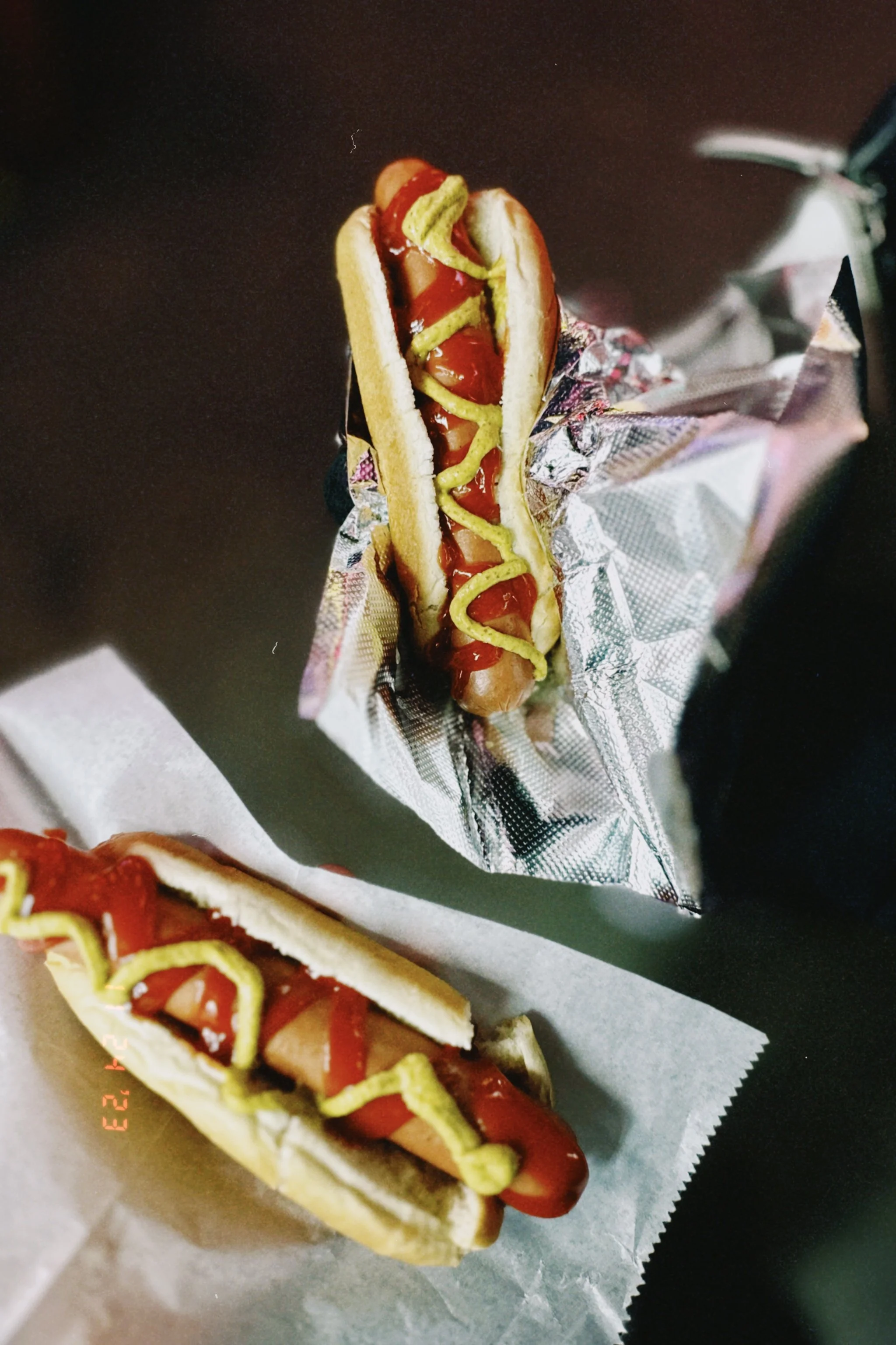 Hot dog topped with ketchup, mustard, and pickle slices, wrapped in foil and placed on a paper tray.