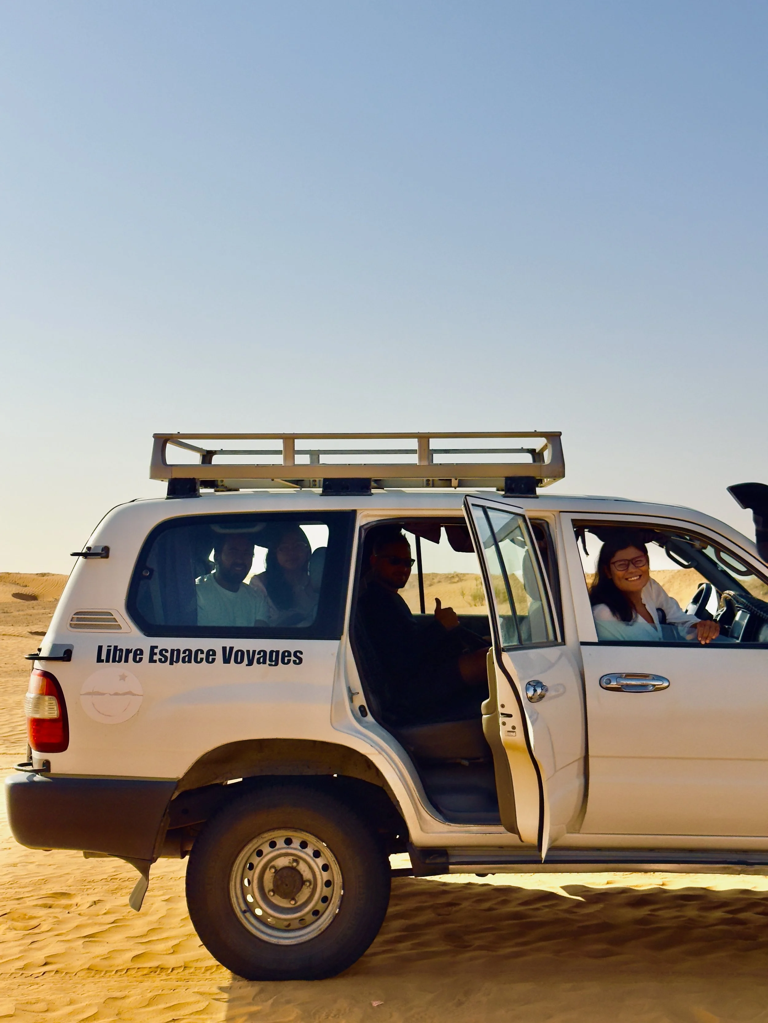 A white SUV with the words "Libre Espace Voyages" on the side, parked on a sandy desert with a few people inside, with a clear blue sky above.