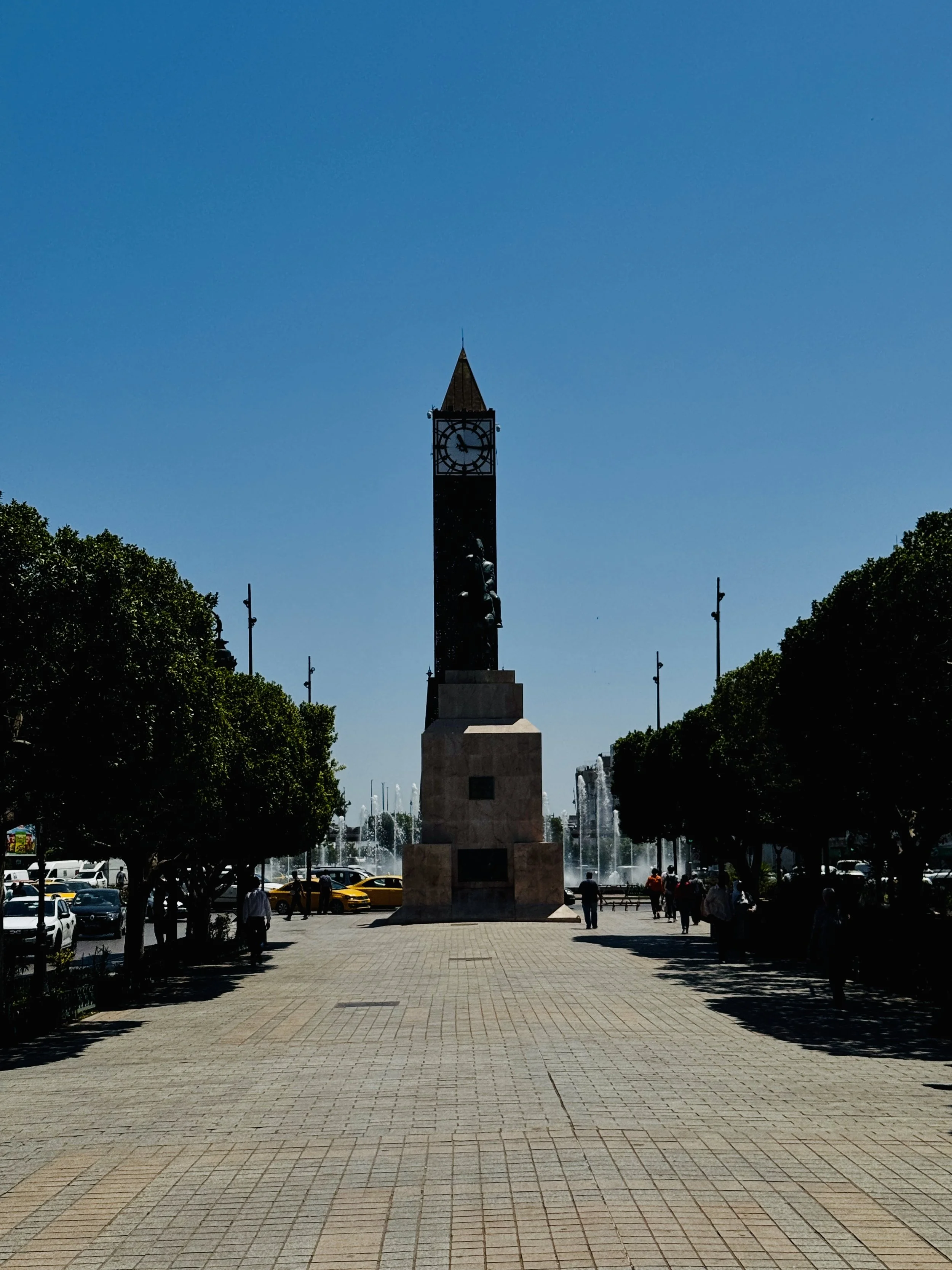 A tall clock tower with a pointed roof, above a fountain with water jets, surrounded by trees and benches, in an urban square.