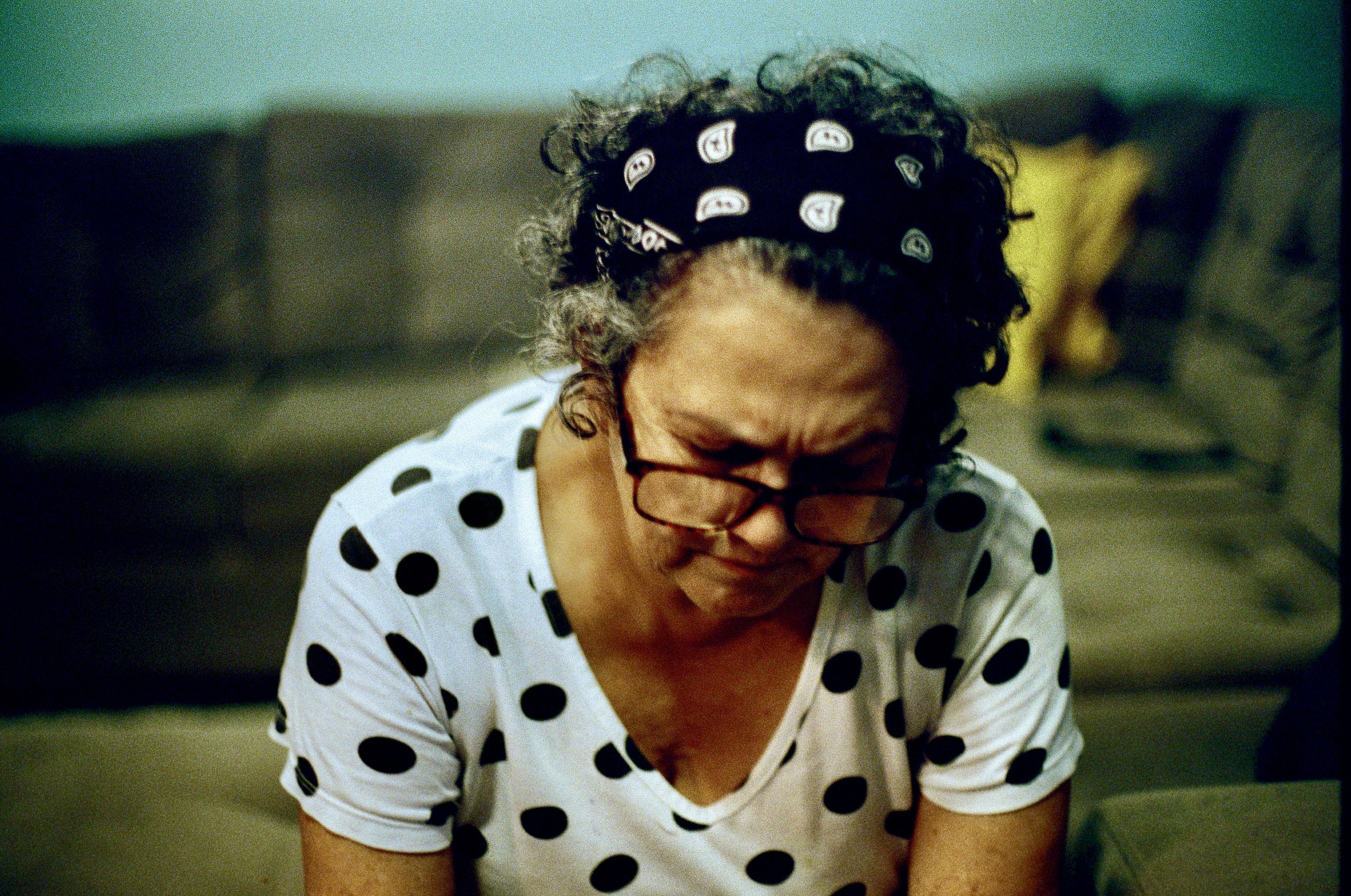 A person with curly hair wearing a black bandana and glasses, wearing a white shirt with black polka dots, looking downward with a focused expression.