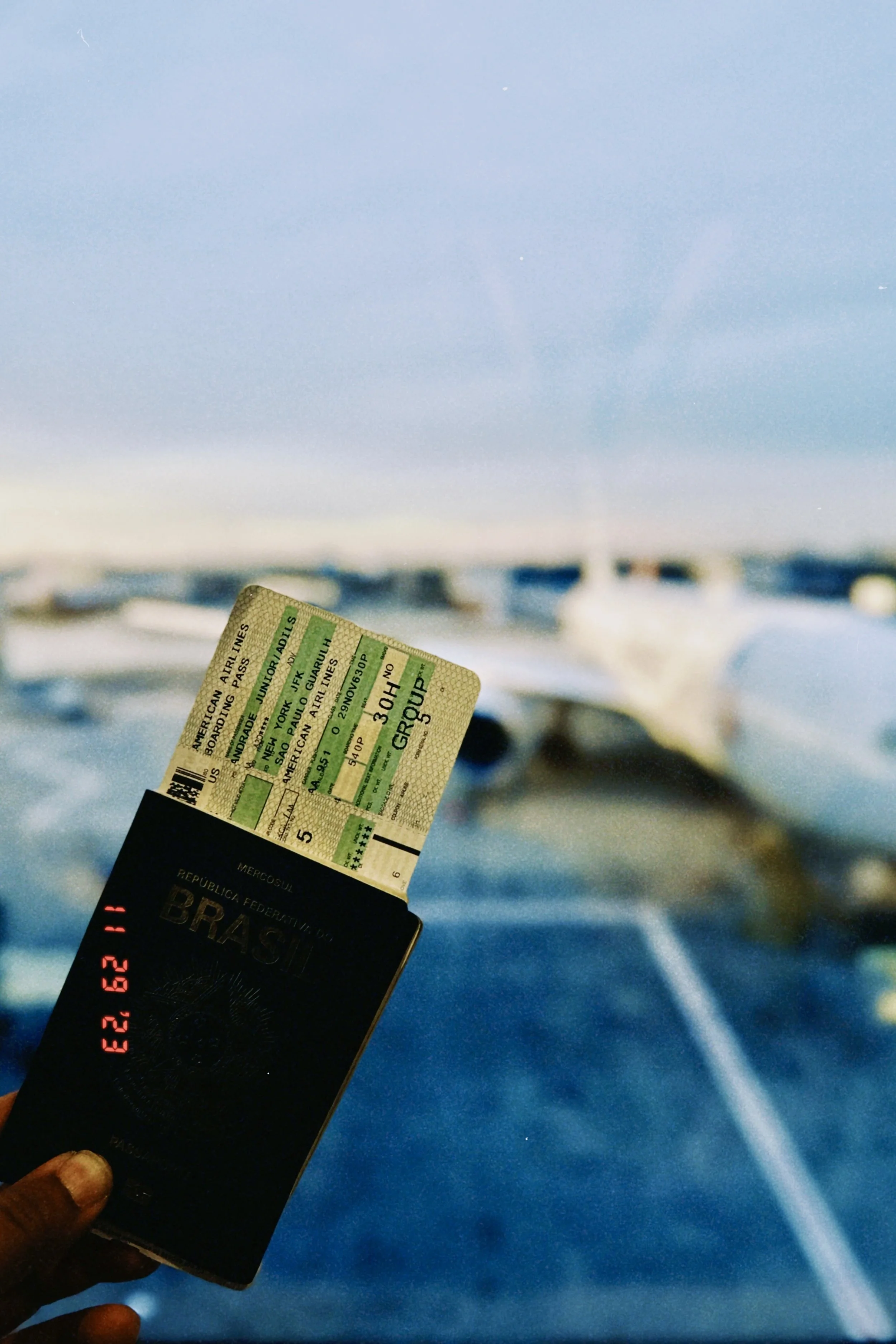 A hand holding a Brazil passport and an American Airlines boarding pass with a blurred airplane window and sky in the background.