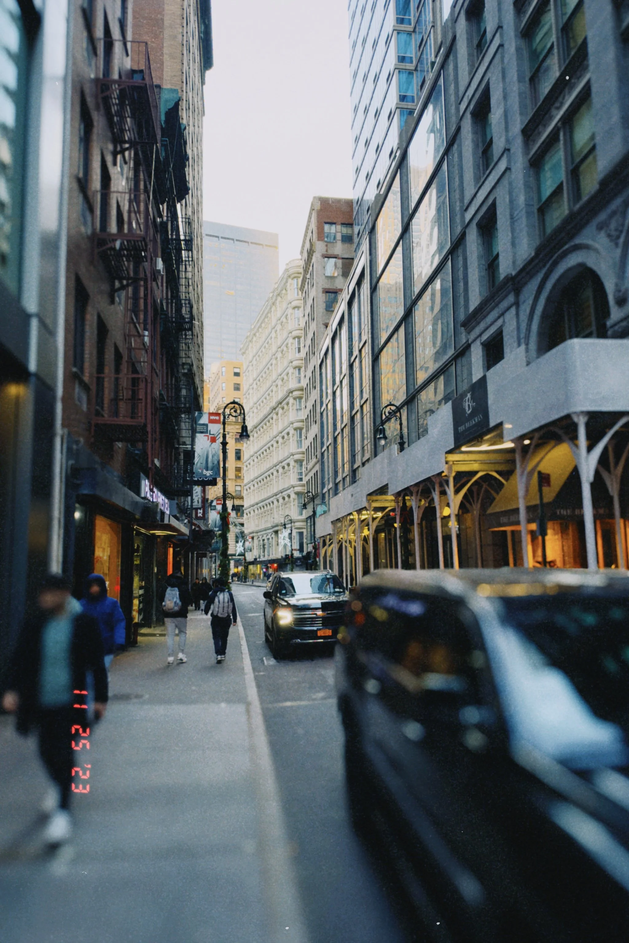 City street with pedestrians walking, cars parked along the curb, tall buildings on both sides, and street lamps.