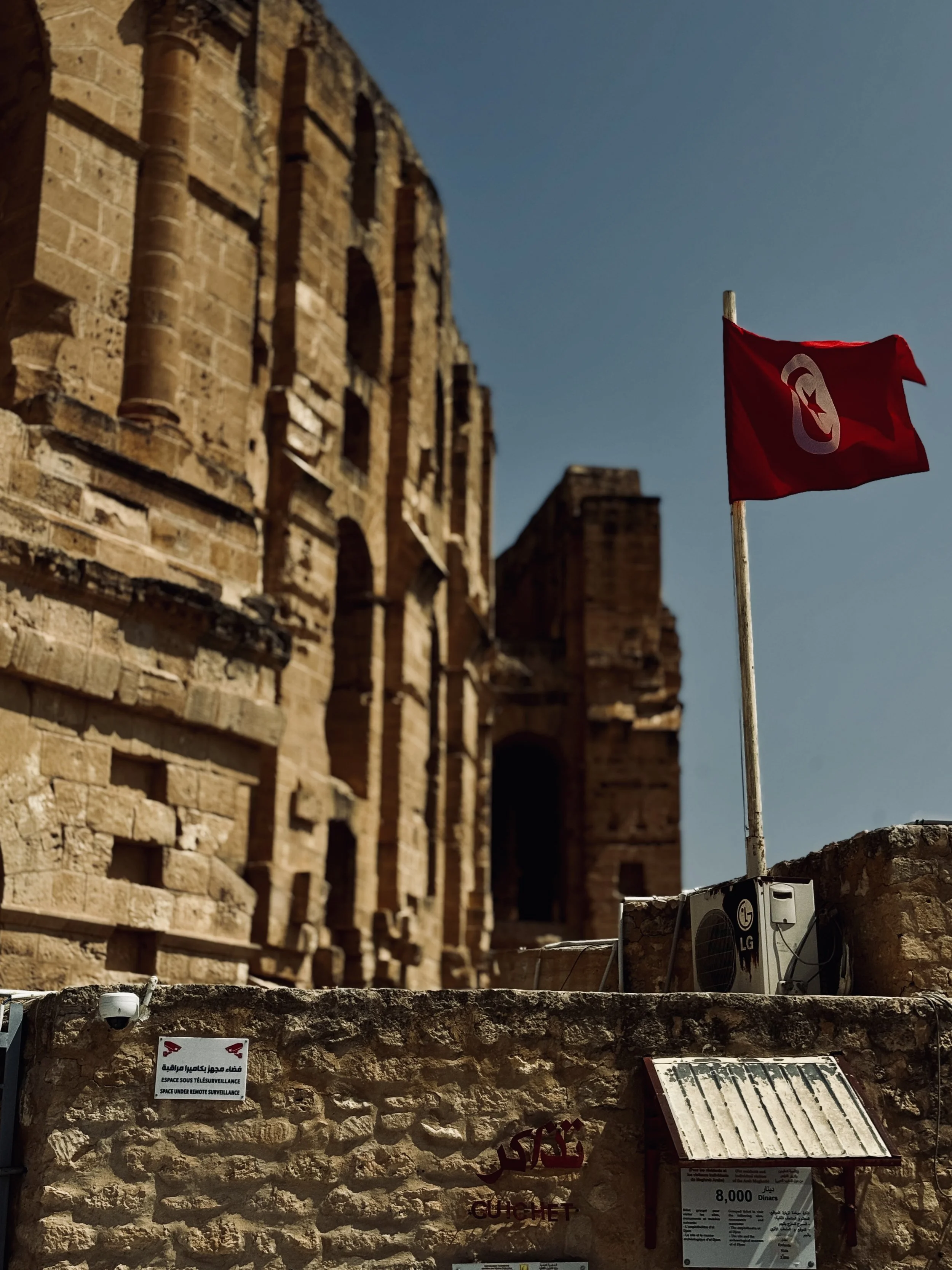 An ancient stone structure with a red flag featuring a white crescent moon and star, situated above modern equipment like an air conditioning unit, with informational signs in multiple languages on the stone wall.