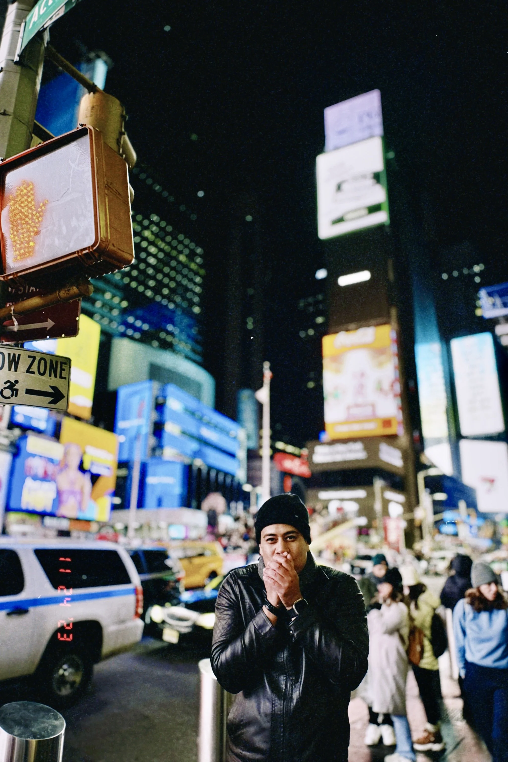 A man in a black jacket and knit cap covering his mouth stands on a busy city street at night, with bright lights, billboards, and crowds in the background.