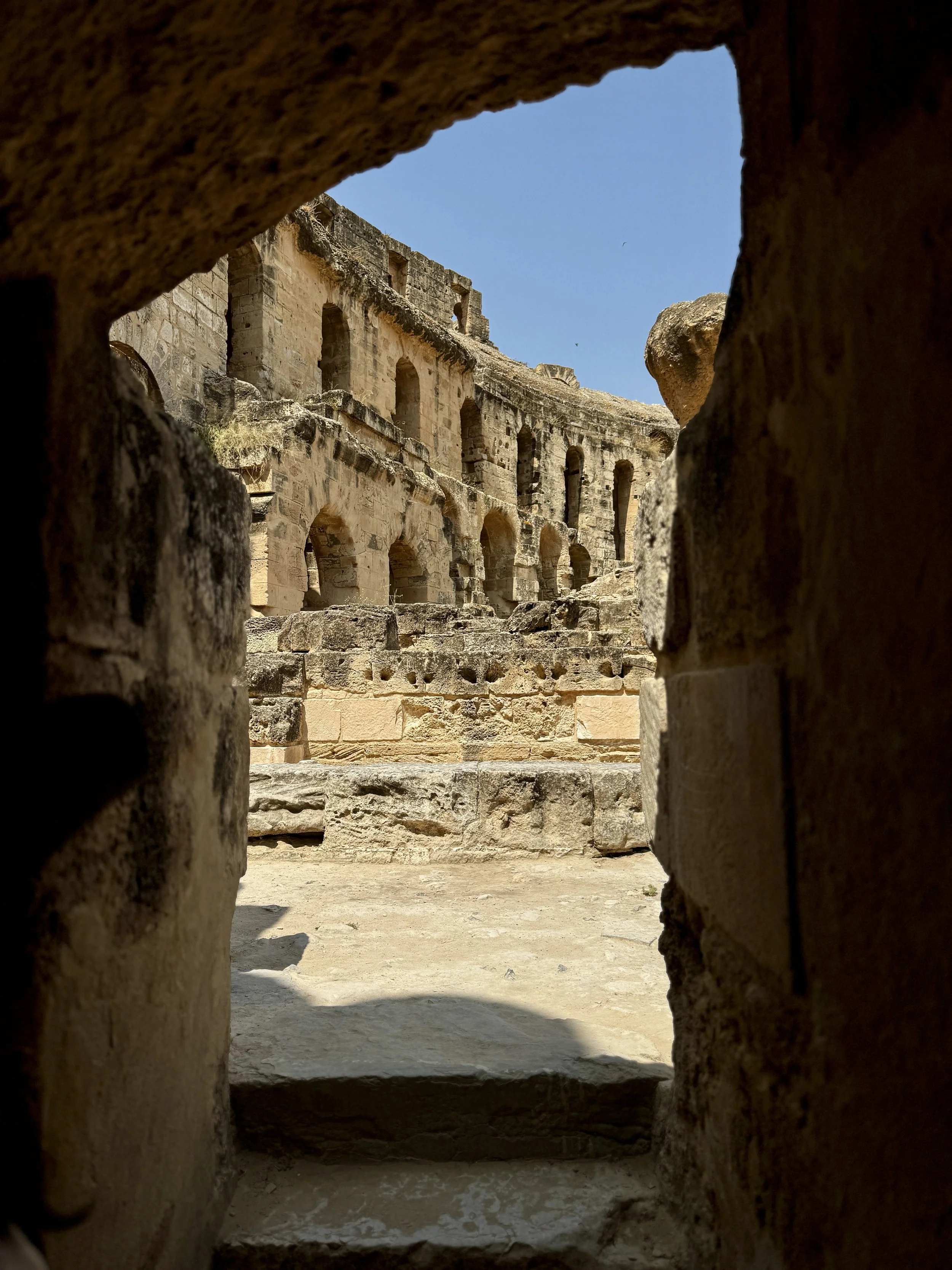Ancient stone Roman amphitheater seen through a small window in a wall, with arches and seating.