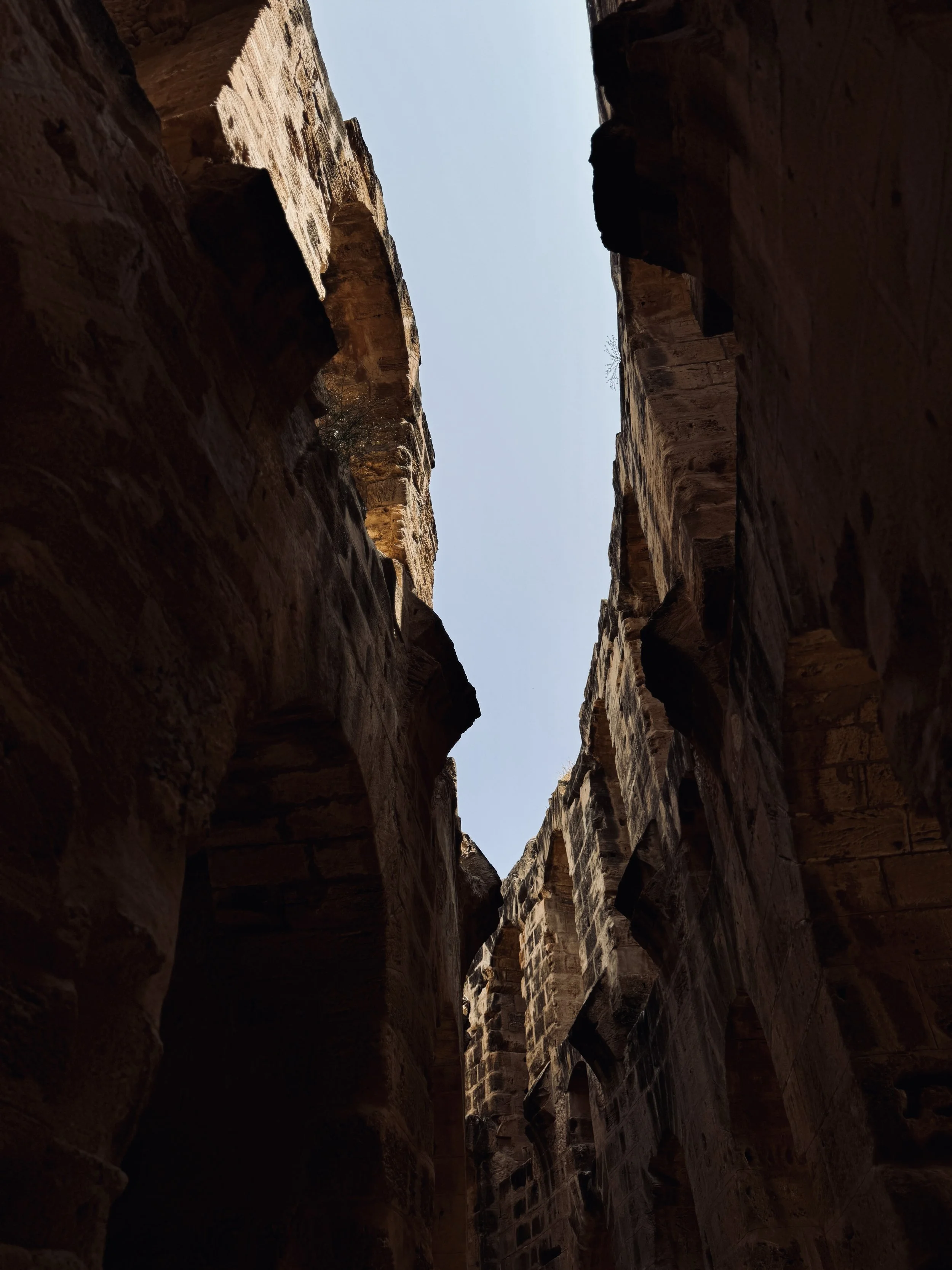 Tall canyon walls with a narrow sky opening at the top, rock formations with layers and shadows, likely in a desert or arid region.