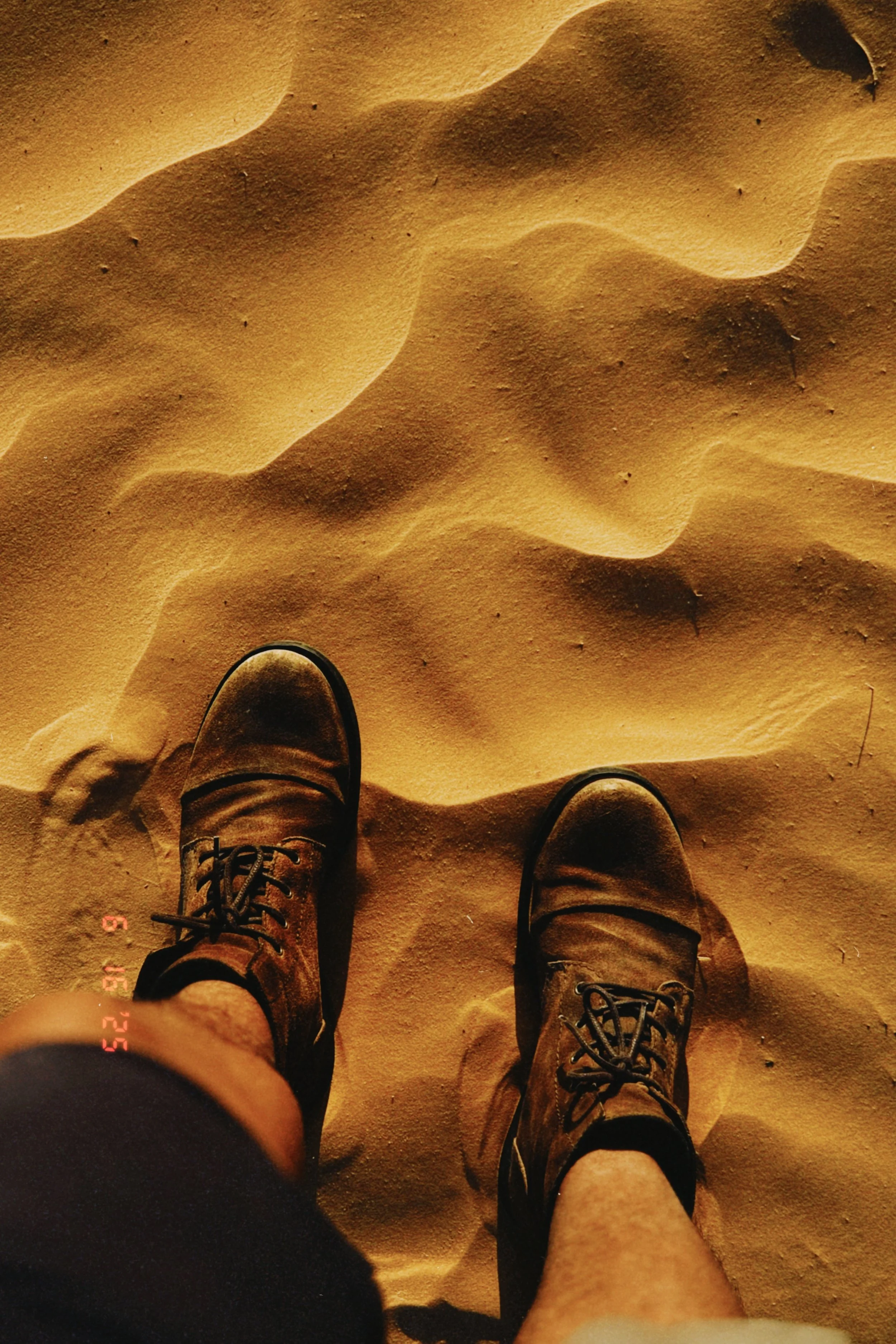 Two people standing on sandy desert terrain, wearing hiking boots, visible from above.