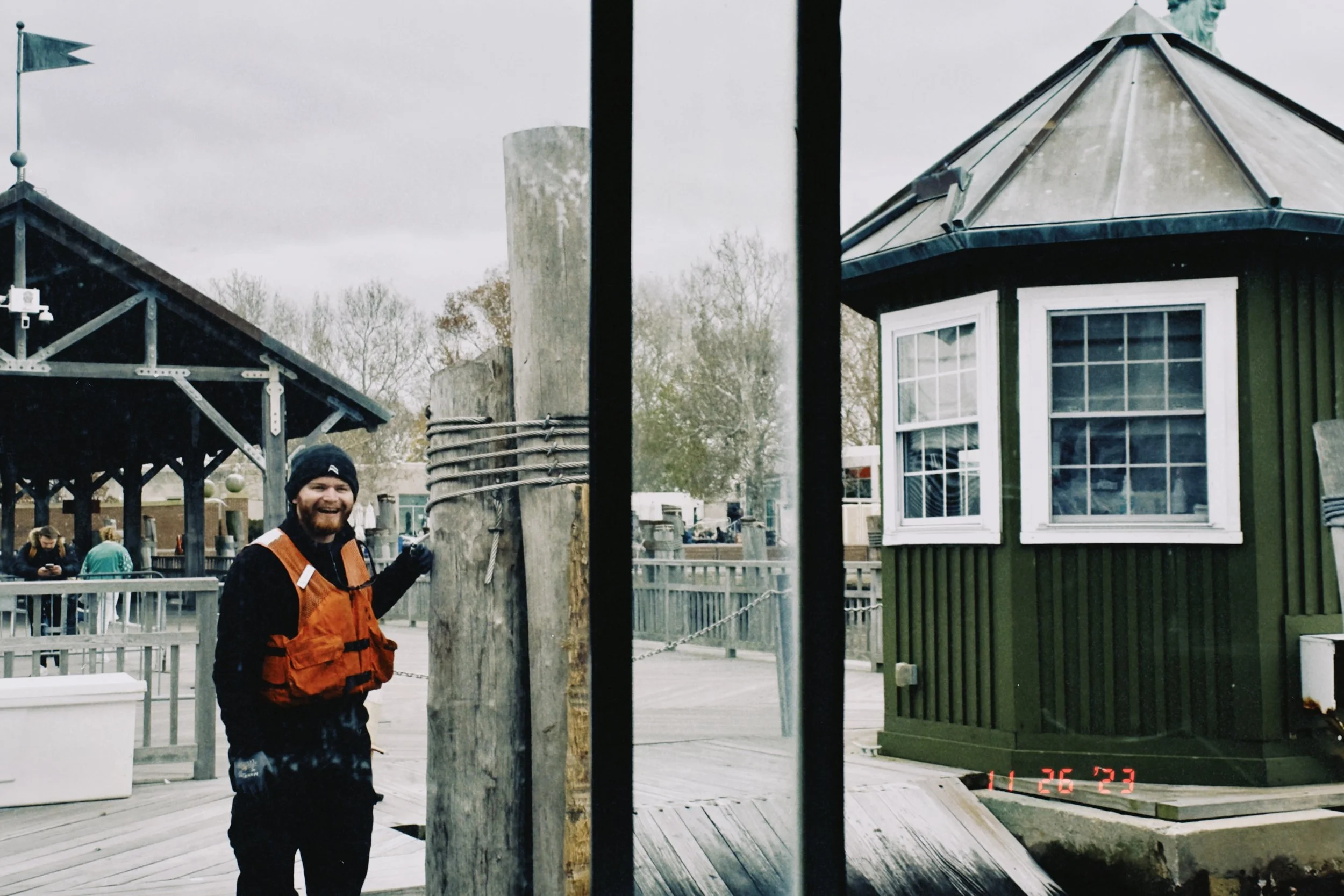 A man with a beard wearing a black beanie and an orange jacket standing outdoors near a wooden utility pole, smiling. There are buildings and trees in the background.
