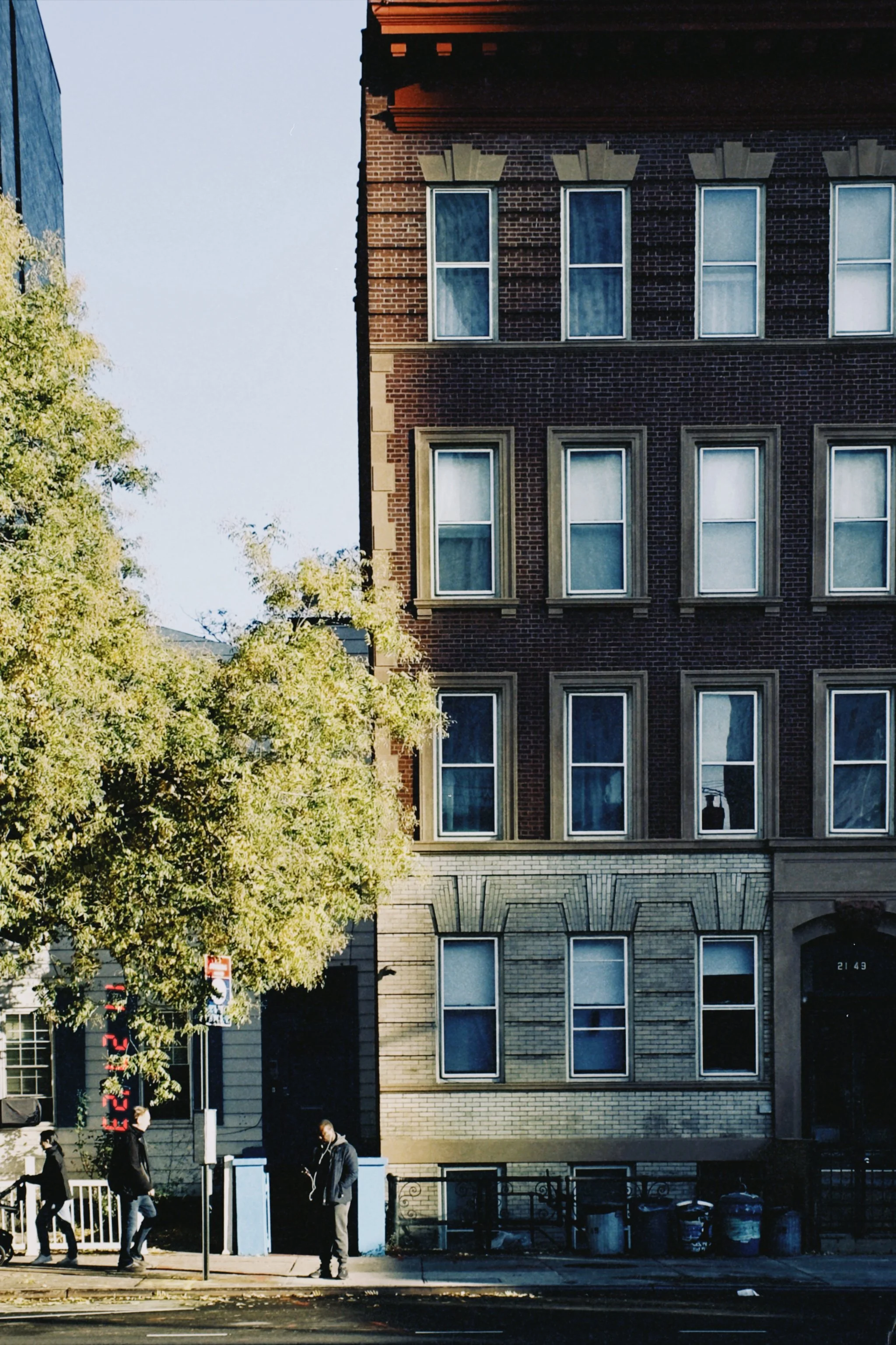 A city street scene with three pedestrians walking on the sidewalk, a green leafy tree, and a multi-story brick building with several windows. There are trash bins and a storefront with digital signage on the ground level.