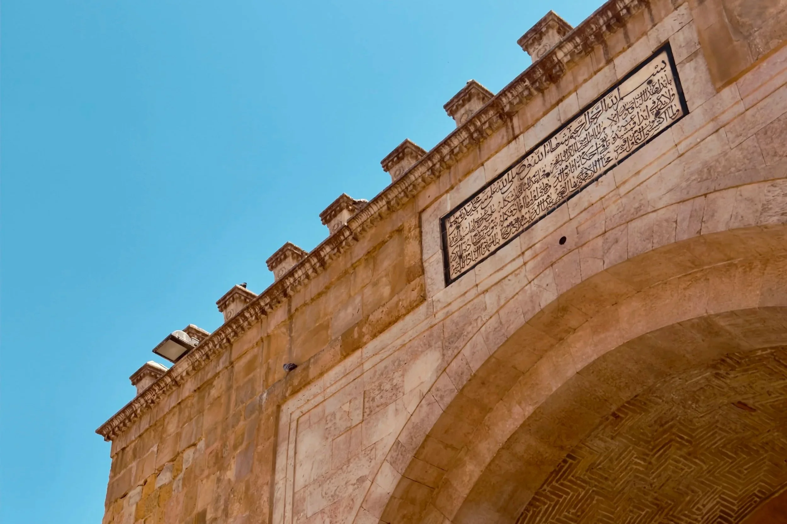 Close-up view of a stone wall with a part of an arch and Arabic inscriptions, under a clear blue sky.