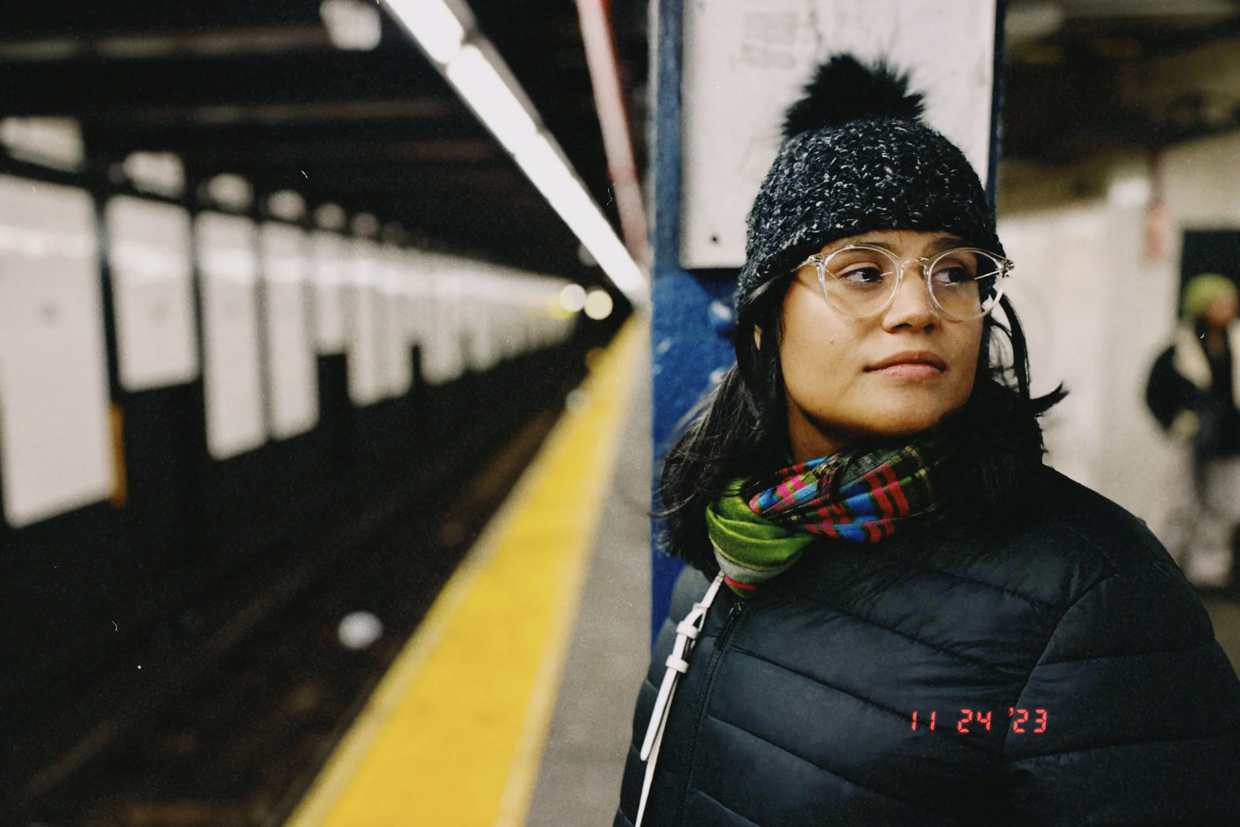 A woman standing on a train platform, wearing glasses, a knit hat with a pom-pom, and a multicolored scarf, looking to the side with a neutral expression.
