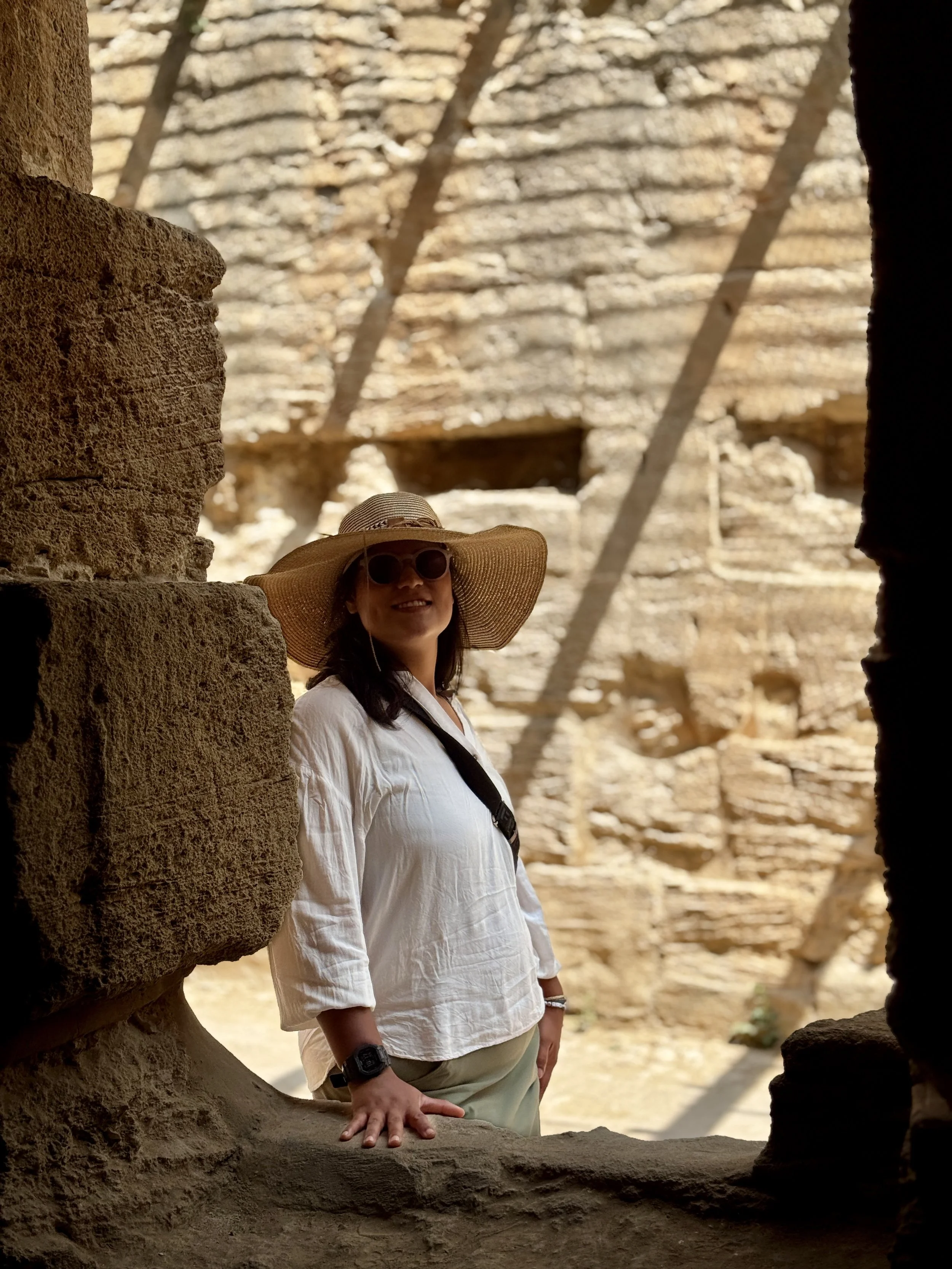 A woman wearing a large sunhat and sunglasses, standing outdoors between stone walls, with a sunny background of rock formations.