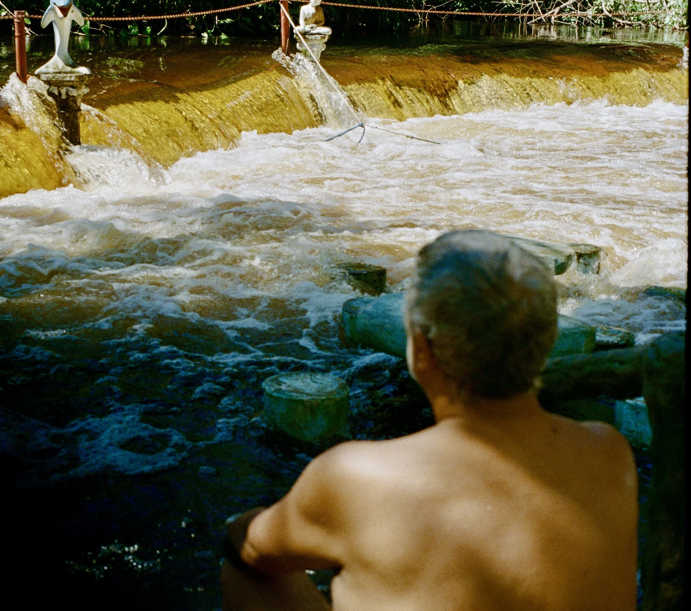 A person with their back to the camera, sitting in a body of water near a waterfall or dam, with water cascading over the edge and rocks in the water.