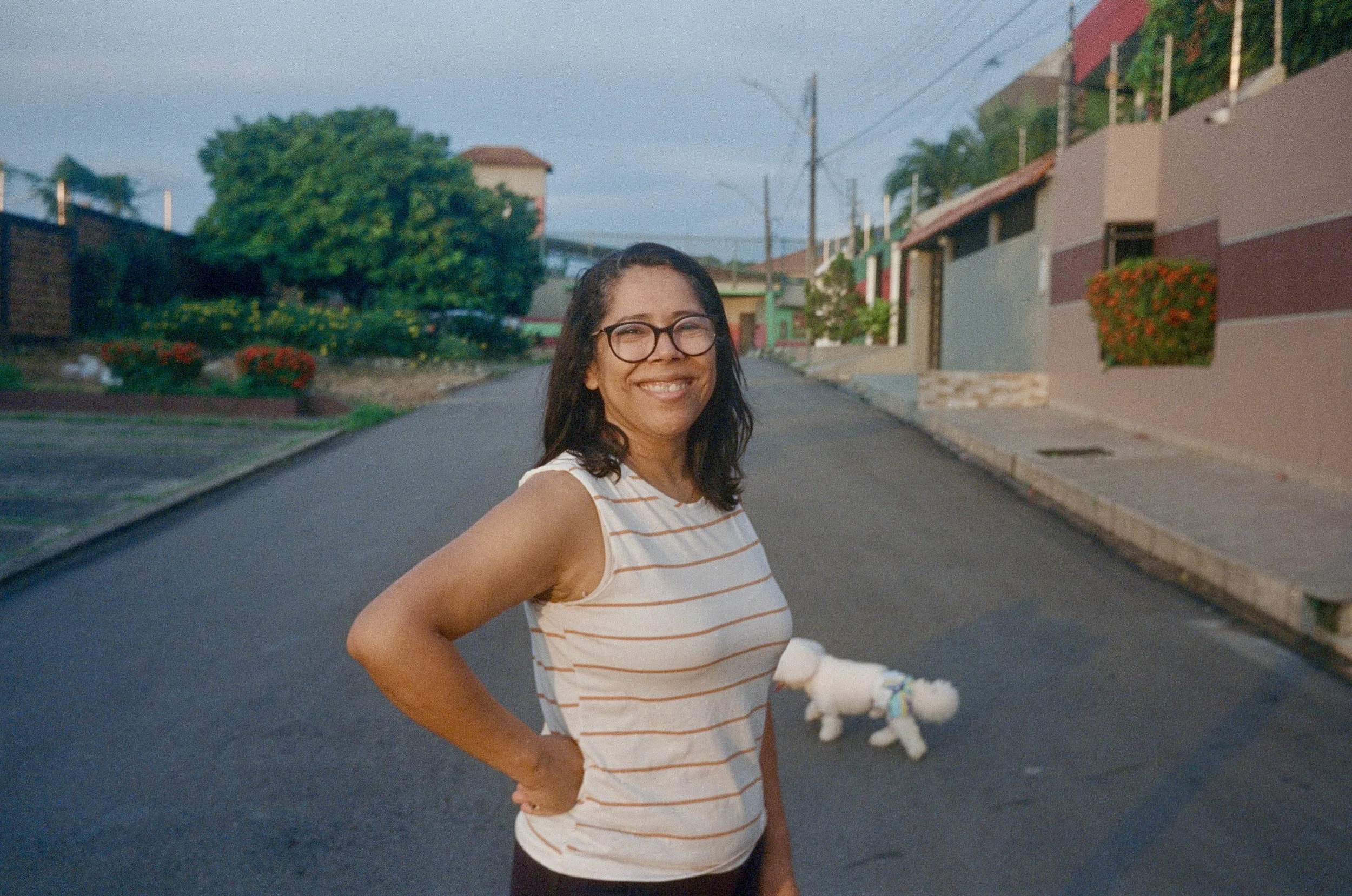 A woman with glasses and a striped sleeveless top smiling and standing on a residential street with a small white dog on a leash in the background.