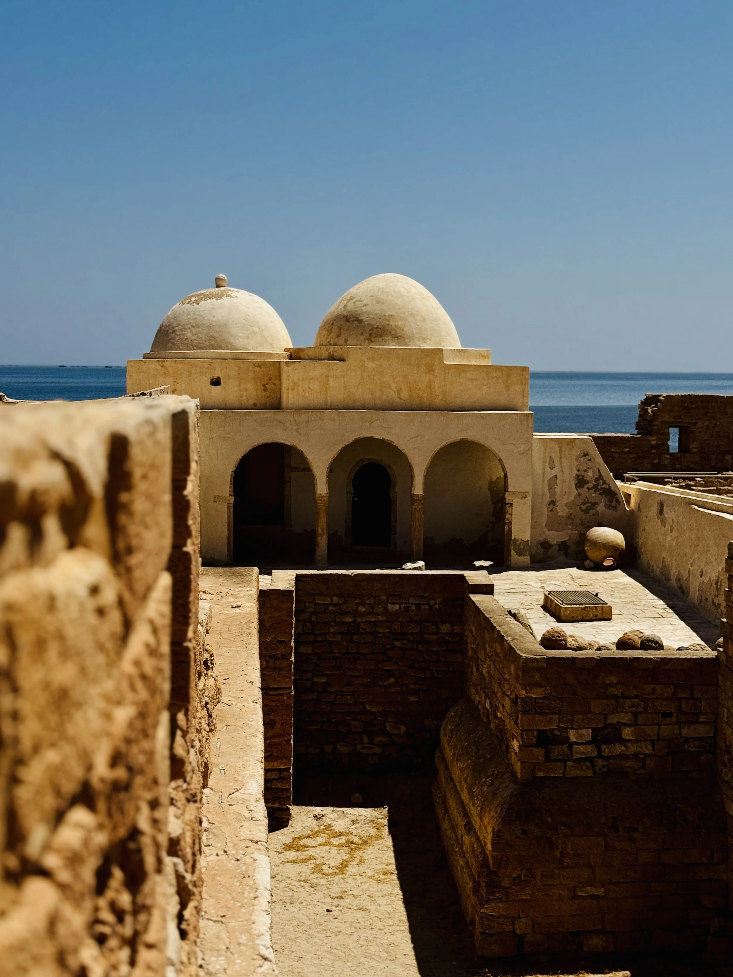 View of two domed structures with arched openings, overlooking the sea under a clear blue sky.