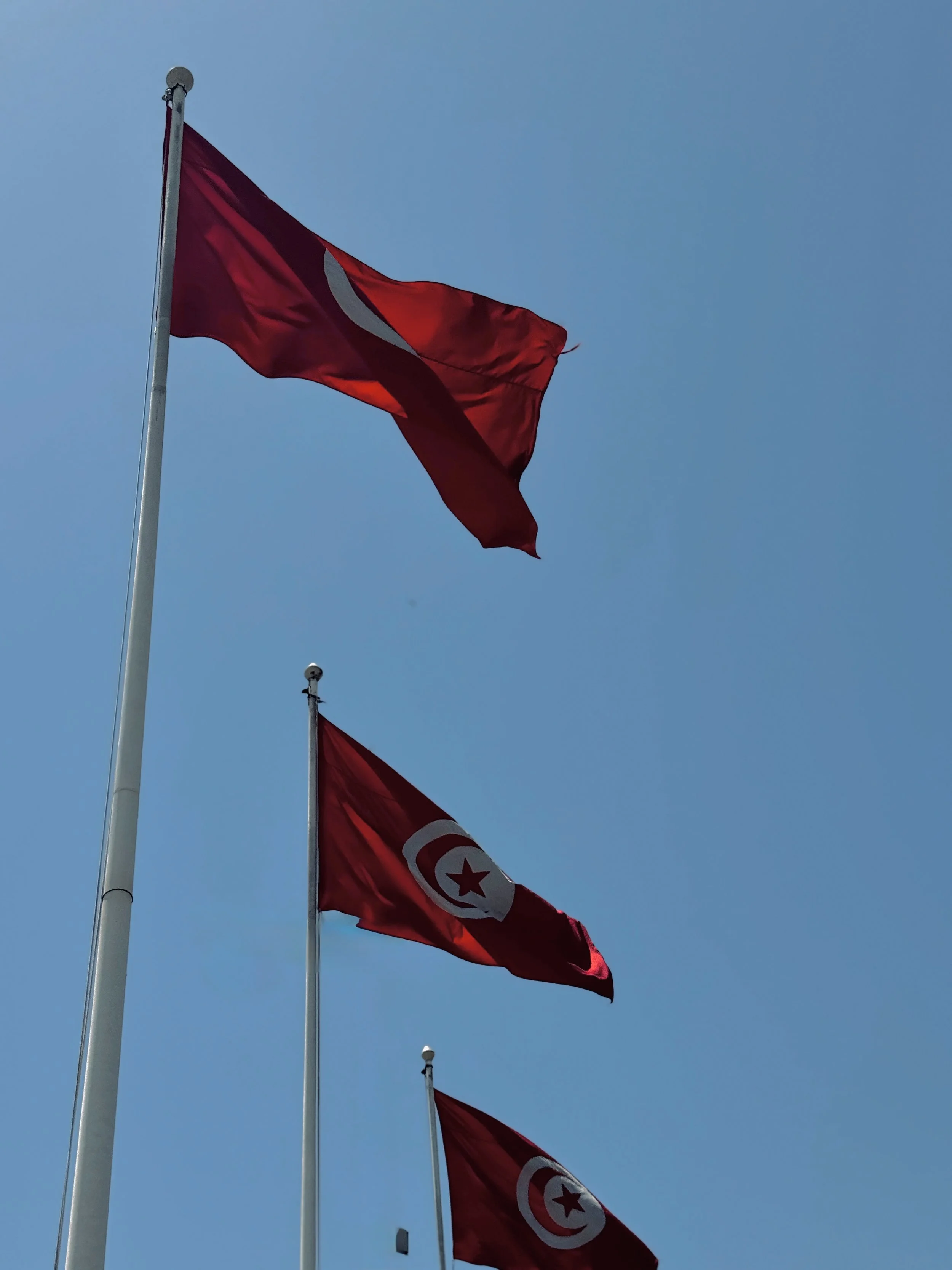 Three flags flying in a clear blue sky, with the top flag being red with a white crescent and star, and the bottom flag partially visible, showing similar colors and symbols.