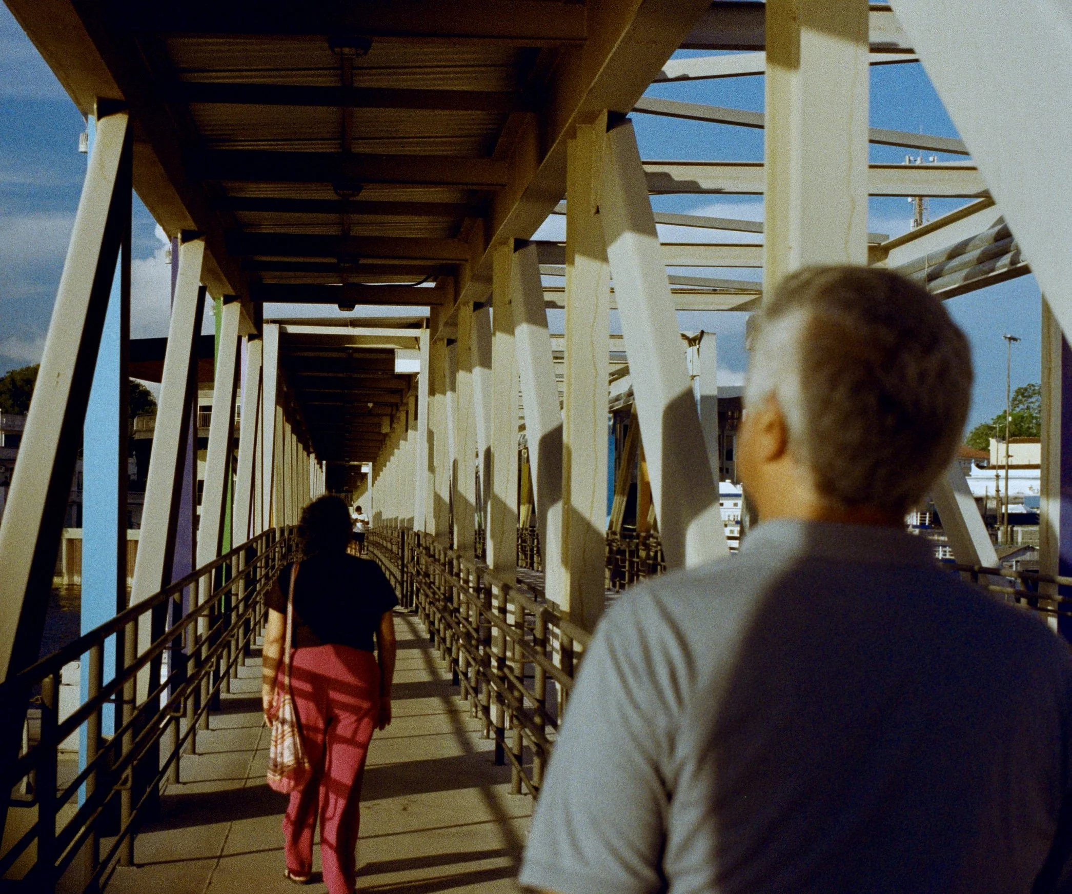 View of a walkway underneath a wooden structure with several people walking, including a person in a black shirt and red pants holding an umbrella, and a child with short hair in the foreground.