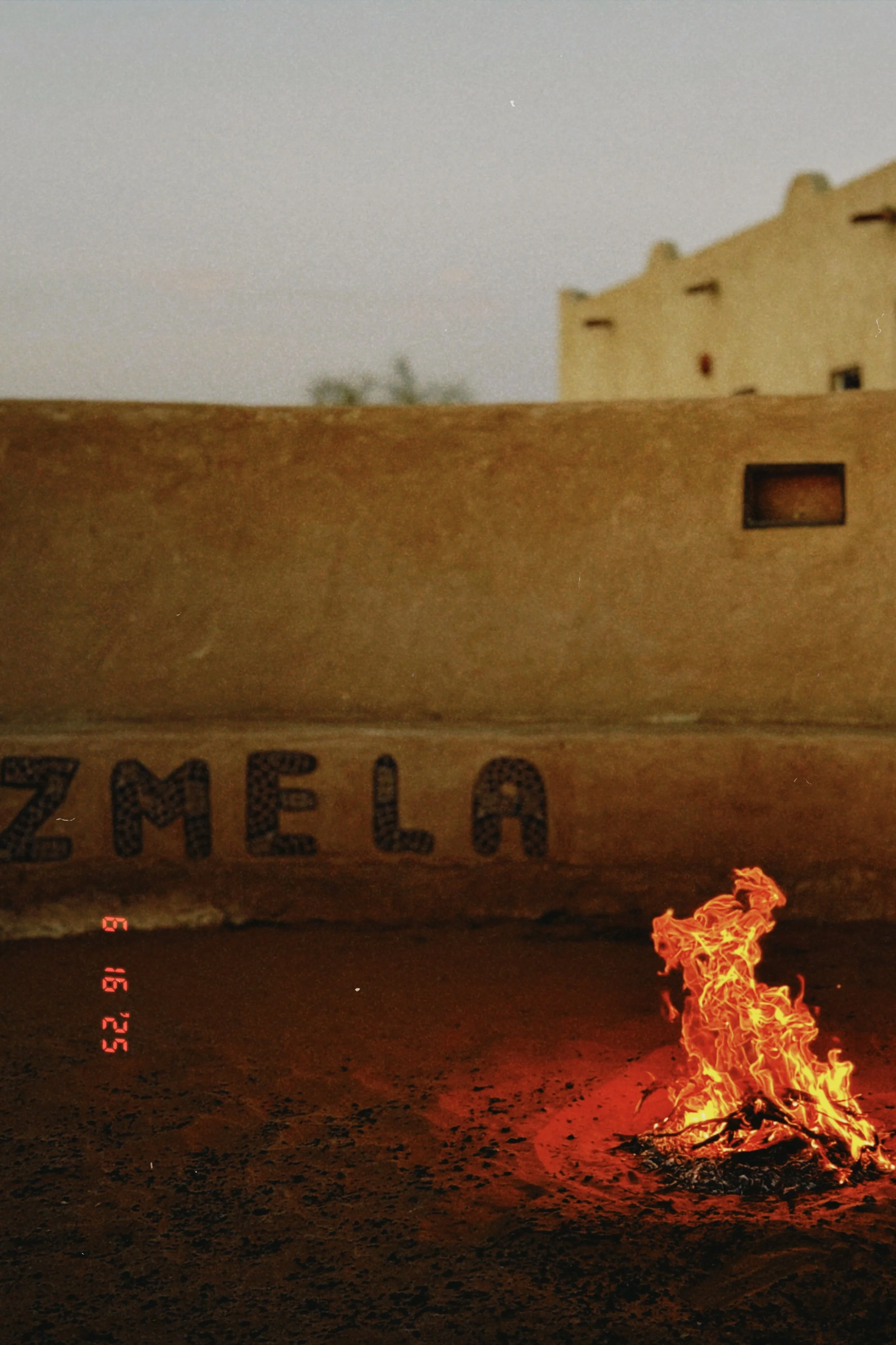 A fire burning on a dirt ground with flames rising, a blurry wall above, and a man-made object with the text 'ZMELEC' partially visible on a cardboard surface.