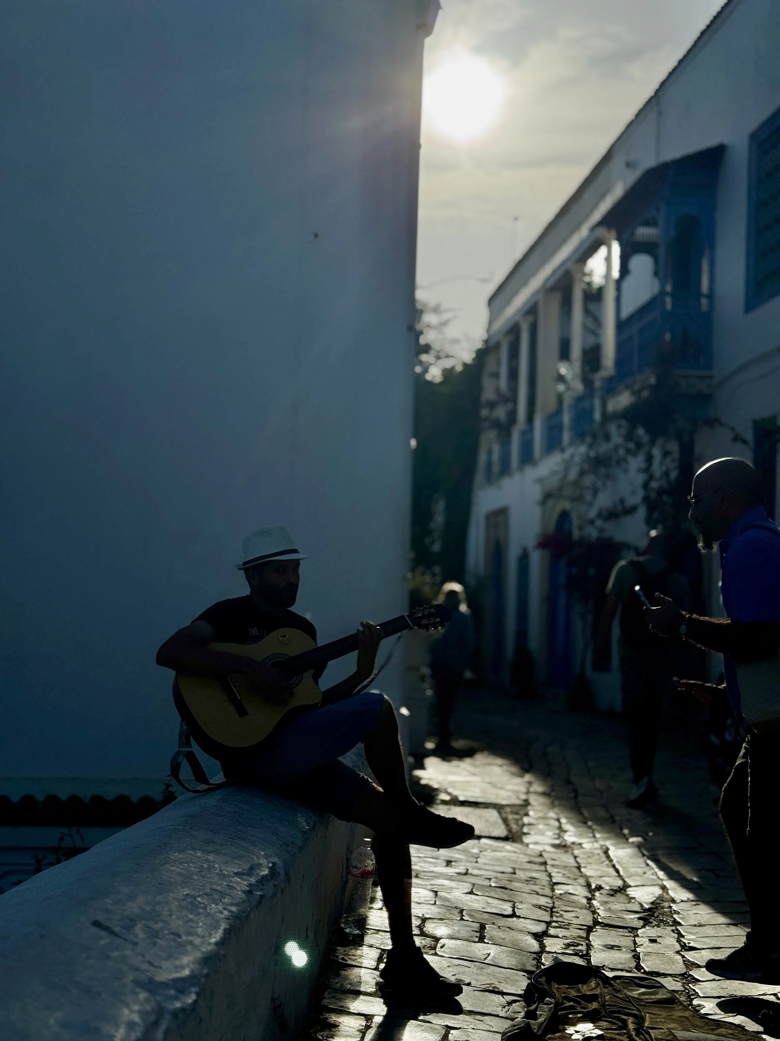 People sitting on a bench in a narrow street, silhouette against the setting sun, with one person playing guitar and others using phones, buildings on both sides, cobblestone street.