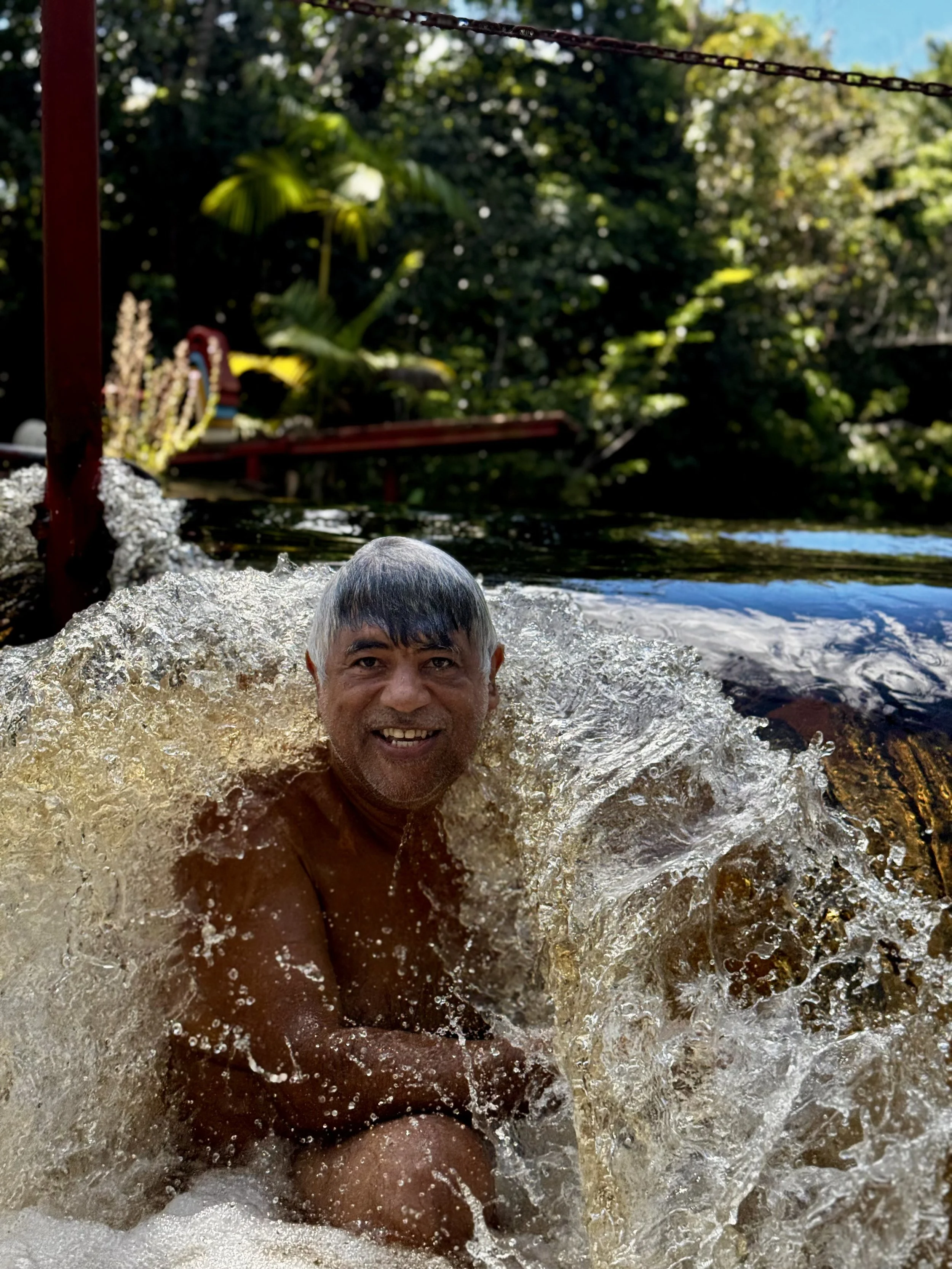 A man with gray hair smiling while swimming in a river or lake with lush green trees in the background.