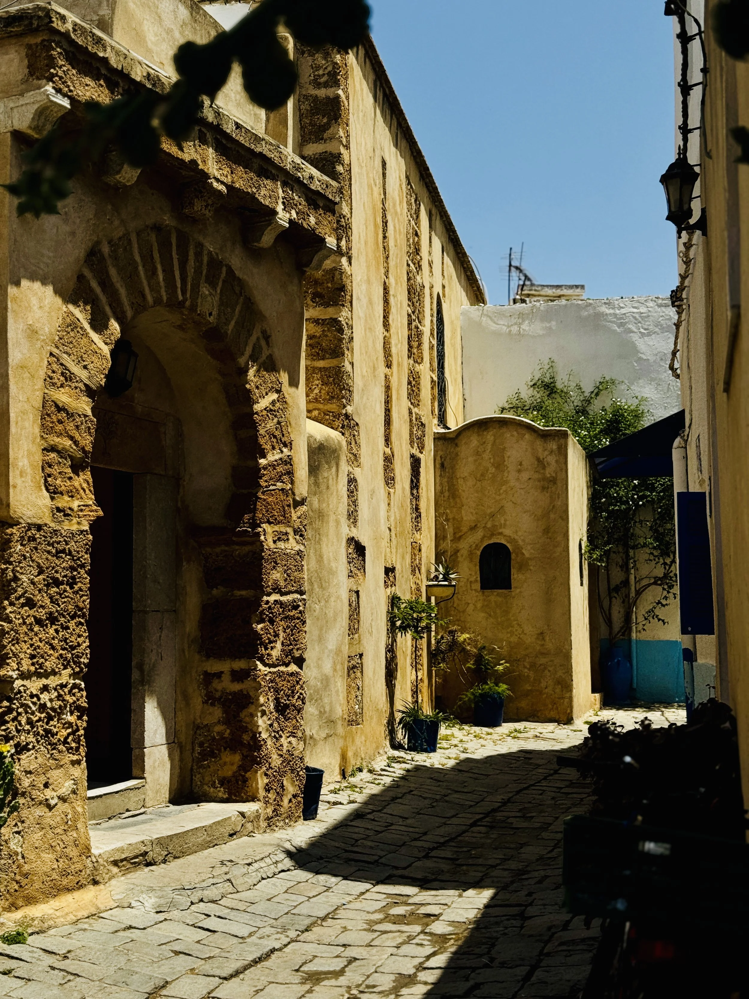 A narrow cobblestone street lined with old stone buildings under a bright blue sky, with a few potted plants and black exterior lamps.