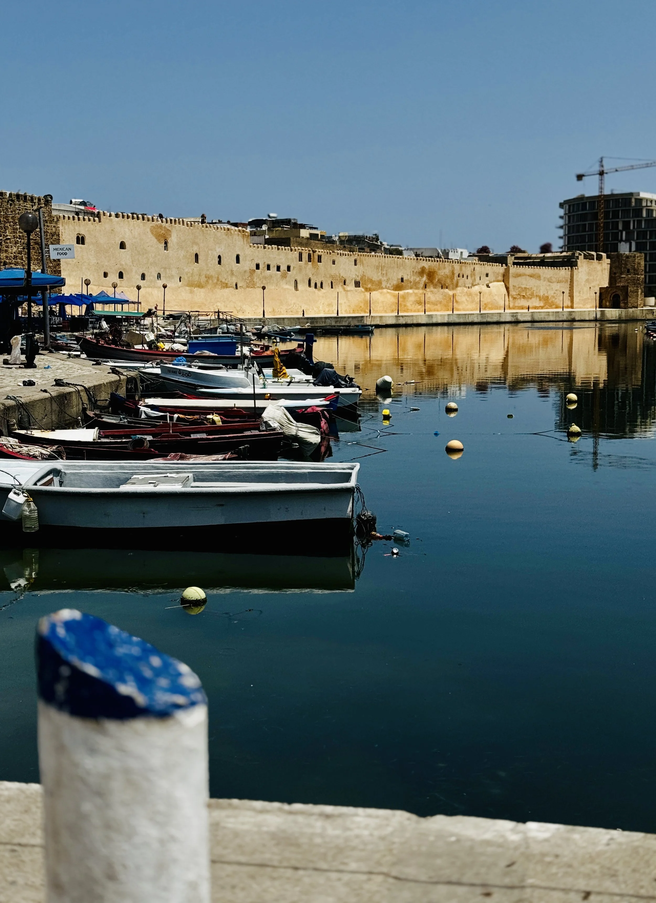 Boats docked in a harbor with a historic stone wall and modern buildings in the background, under a clear blue sky.