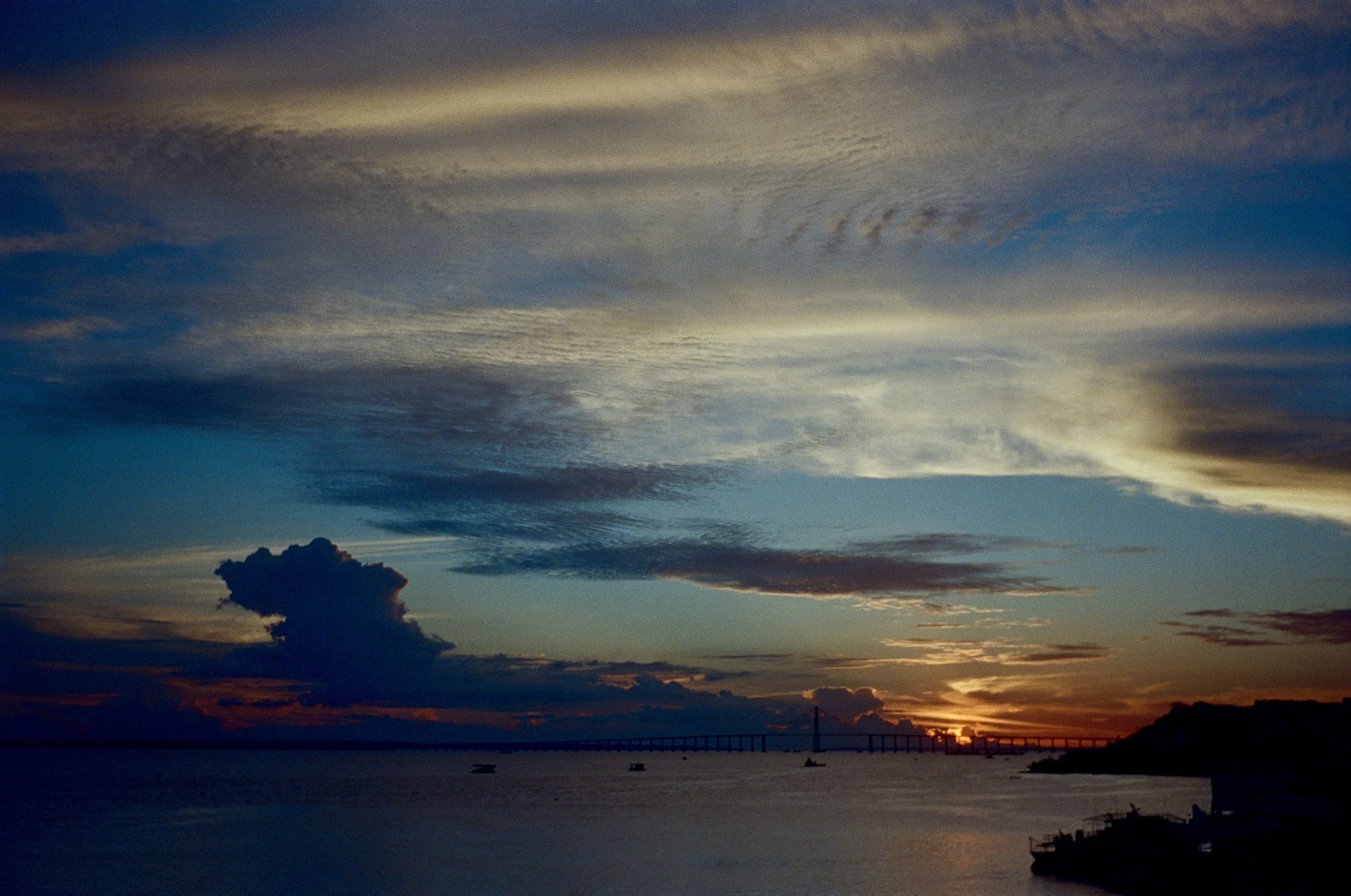 Sunset over a body of water with a bridge in the distance, dark clouds in the sky, and silhouettes of trees and boats along the shore.