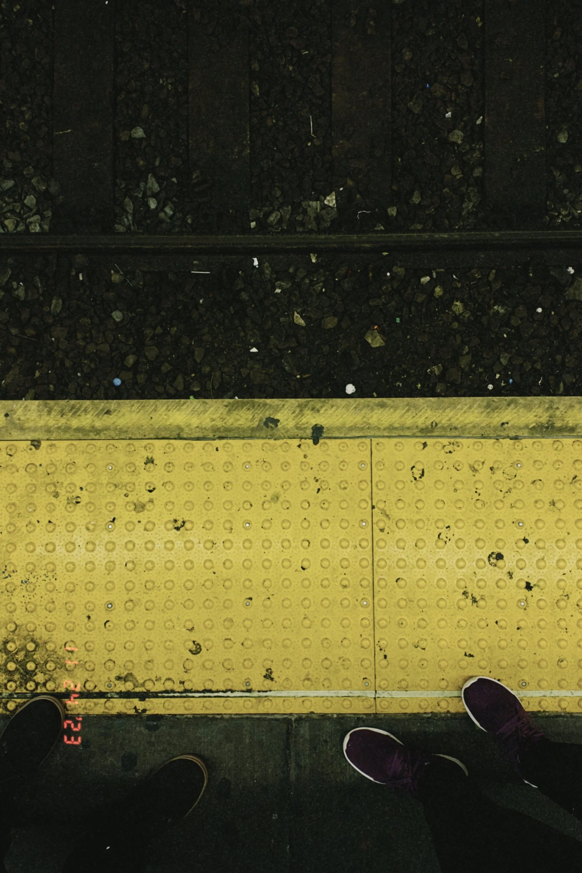 View of a train platform with two pairs of shoes visible in the bottom corners, yellow tactile paving in the middle, and dark train tracks in the background.